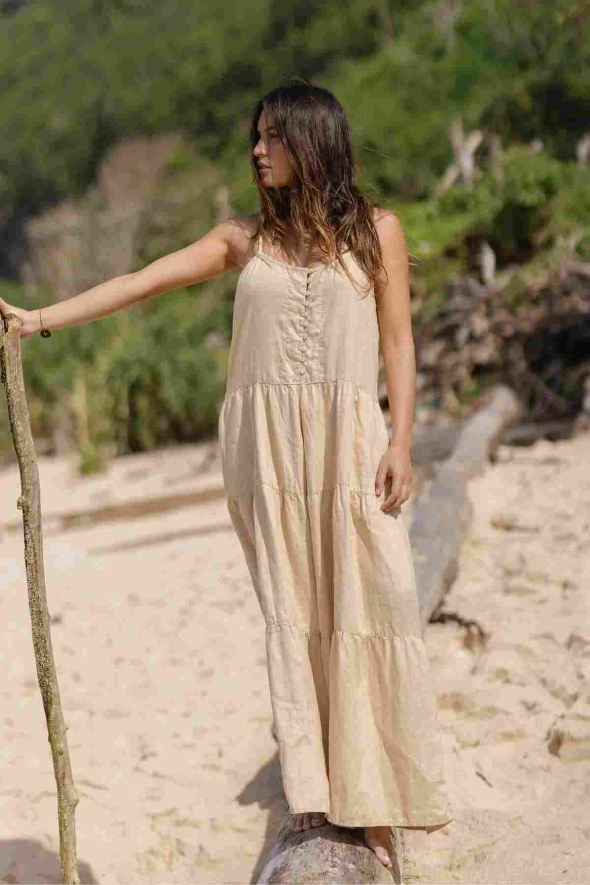 A woman in a made-to-order La Mexicana Playsuit is standing on the sandy beach, her attire crafted from 100% linen by Myrah Penaloza. She holds a stick and gazes to the side as lush green foliage and a large log on the sand complement her serene summer moment.