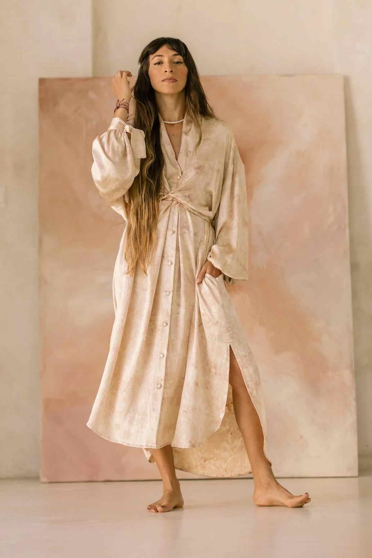 A woman with long hair stands barefoot indoors, wearing a light, flowy Silk Inanna Wrap Gown by Myrah Penaloza, made of botanically hand-dyed silk with a slit. Her left hand rests on her head, and she has accessories on her right wrist. The background features a soft, pinkish-beige abstract backdrop created by artisan tailors.