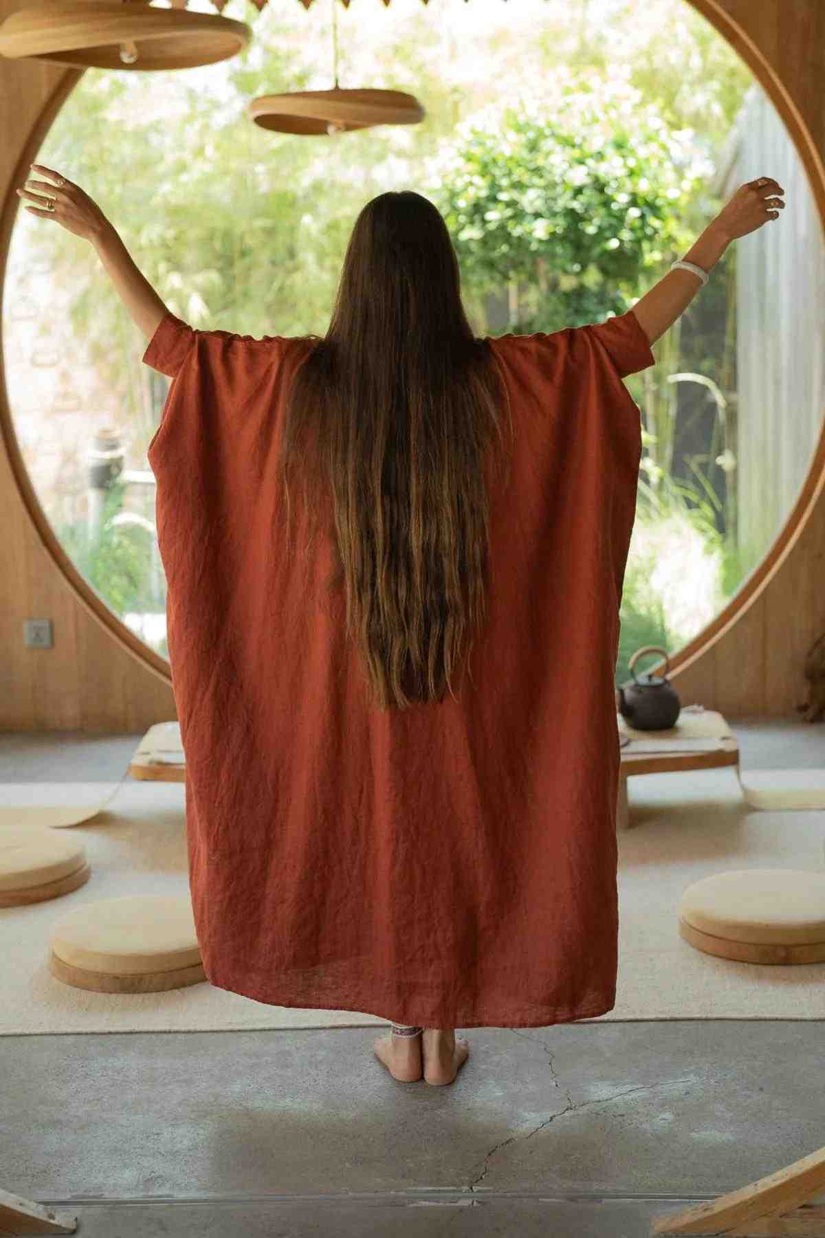 A person with long hair stands with arms raised, gracefully draped in a flowing La Majia Kaftan by Myrah Penaloza. They are inside a room featuring round windows and standing in front of a large circular window. The space is adorned with minimalistic wooden decor and round cushions on the floor, evoking the artistic spirit of Frida Kahlo.