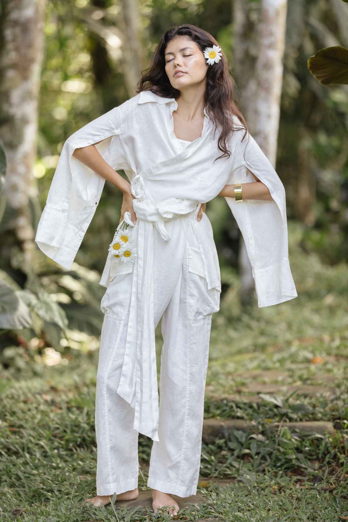 A person stands outdoors, donning the versatile and stylish Sutra Linen Set by Myrah Penaloza, a made-to-order loose-fitting, white ensemble featuring flared sleeves and wide legs. They have a flower tucked behind their ear and one in their pocket. Their dark hair cascades down against a lush green background with blurred foliage.