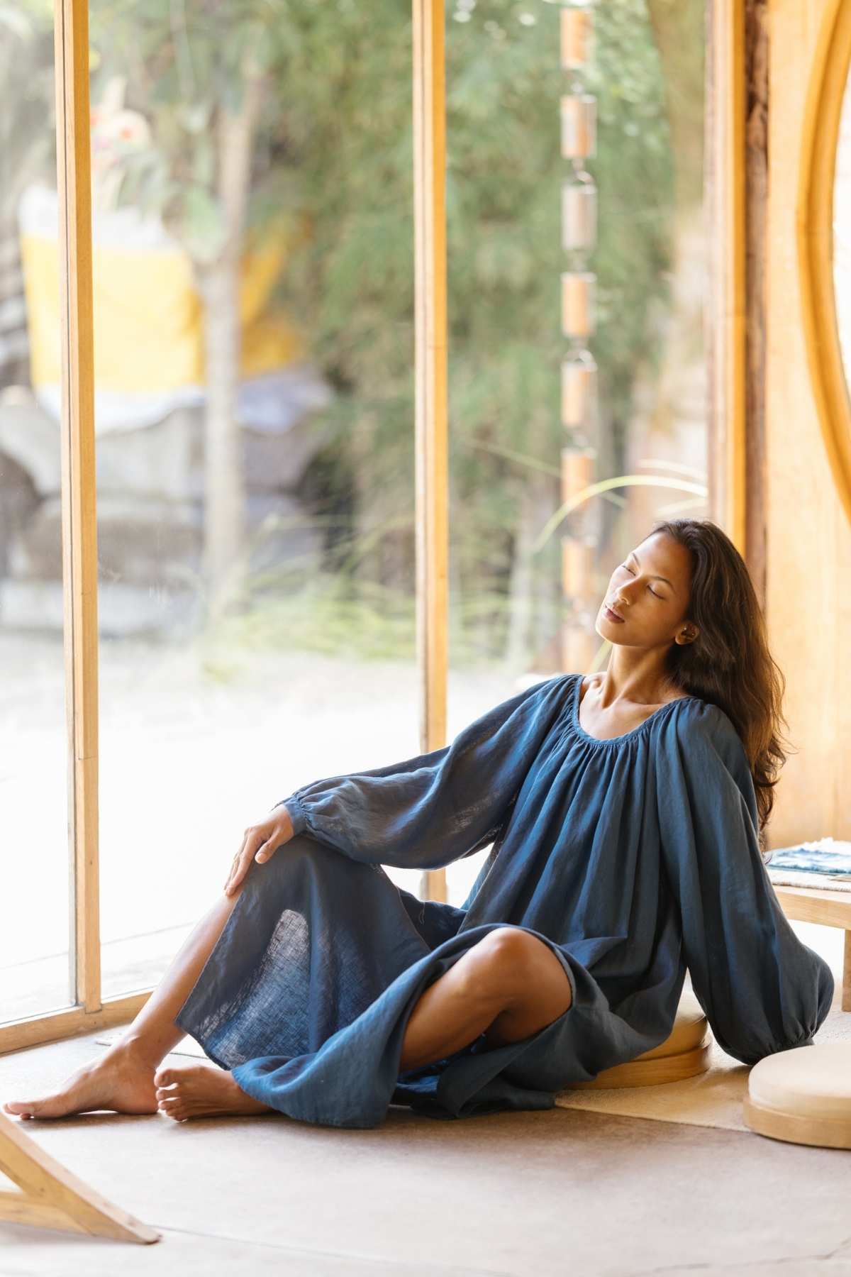 A person with long hair, dressed in a flowing Alchemy Gown (Crinkle Linen Cotton) by Myrah Penaloza, sits relaxed on the floor by a large window, basking in sunlight. The background features a serene outdoor scene with green foliage. The individual appears calm and peaceful, with eyes closed and a slight smile, embodying the essence of a modern mystic.