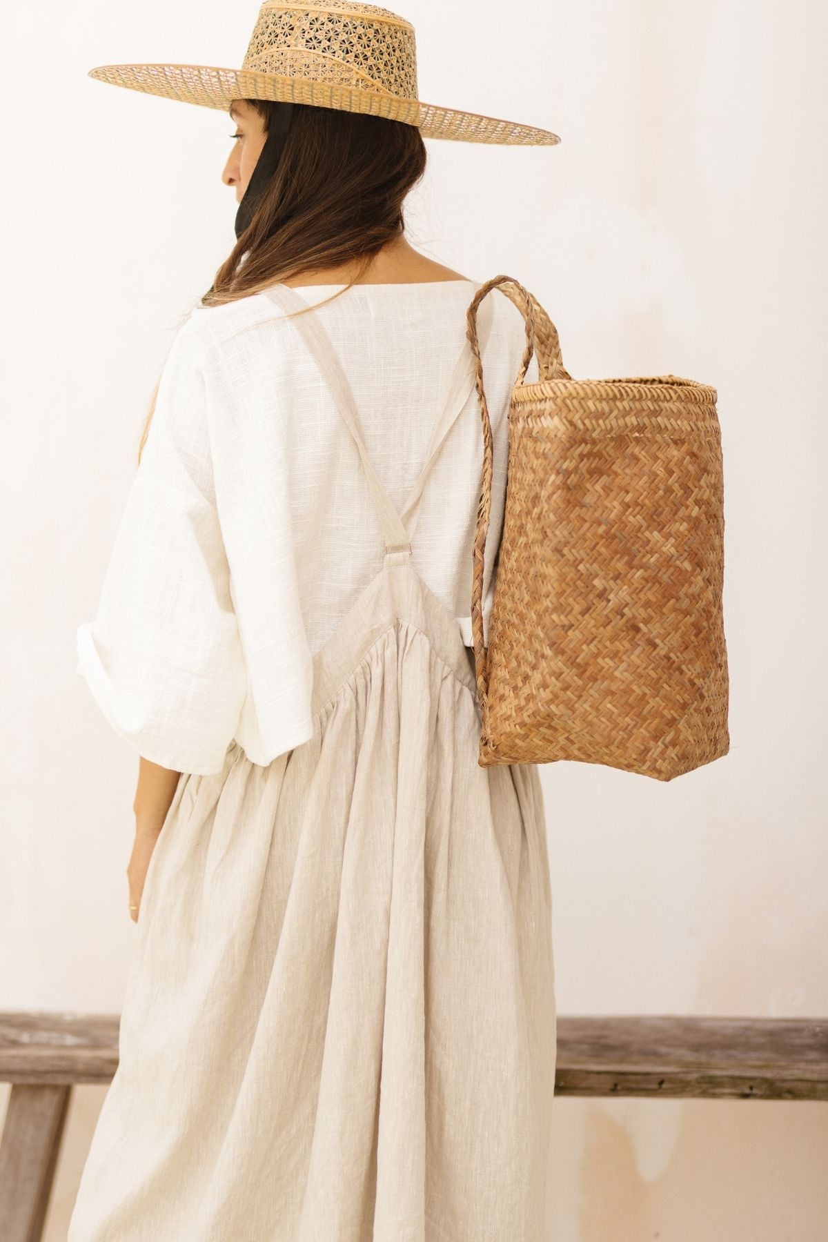 A woman wearing the May May Overall Skirt from Myrah Penaloza stands with her back to the camera. She also wears a straw hat and carries a large woven basket backpack. The background is neutral and uncluttered, highlighting her relaxed, summery outfit.