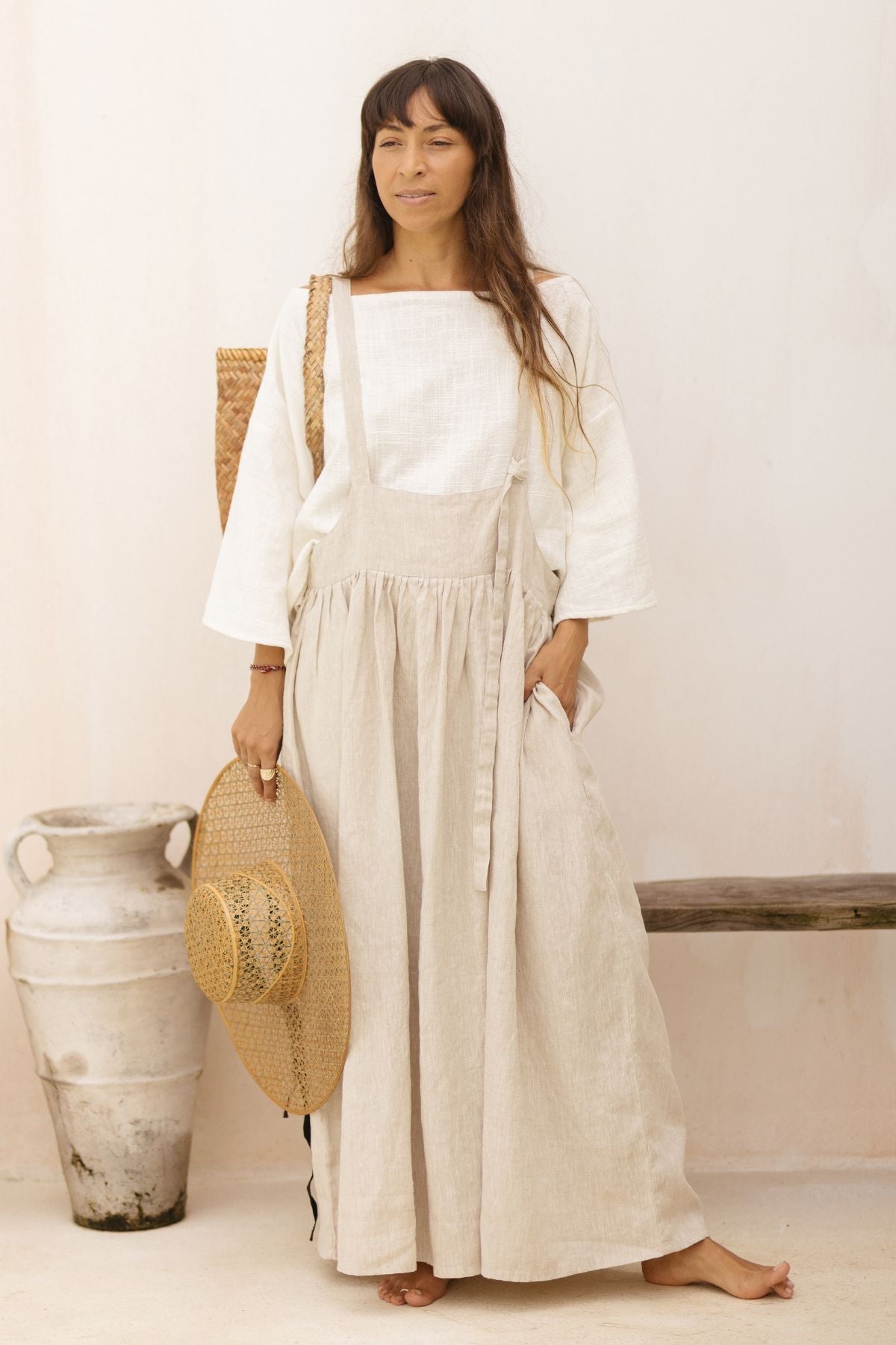 A woman stands barefoot indoors against a light-colored wall, wearing the May May Overall Skirt by Myrah Penaloza, paired with a white blouse underneath. She holds a wide-brimmed straw hat in one hand and has a woven bag over her shoulder. A large clay vase and a wooden bench are beside her.