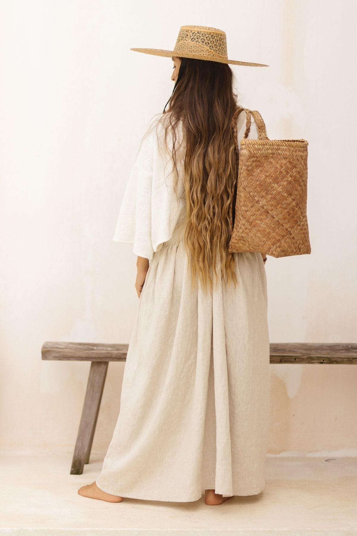 A person with long wavy hair stands barefoot, facing away from the camera. They wear a wide-brimmed straw hat, a white blouse, and a light-colored May May Overall Skirt made of crinkle linen fabric by Myrah Penaloza. They carry a large, woven straw bag on their shoulder. A wooden bench adds charm to the magical fairy garden in the background.