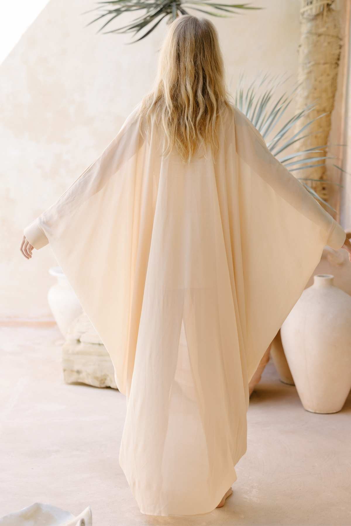 A person with long, wavy hair, photographed from behind, wears a flowing Leo Kaftan Chiffon Sheer by Myrah Penaloza made from sheer silk with wide sleeves. They stand in a softly lit room adorned with large, pale pottery and some greenery in the background, creating a serene atmosphere.