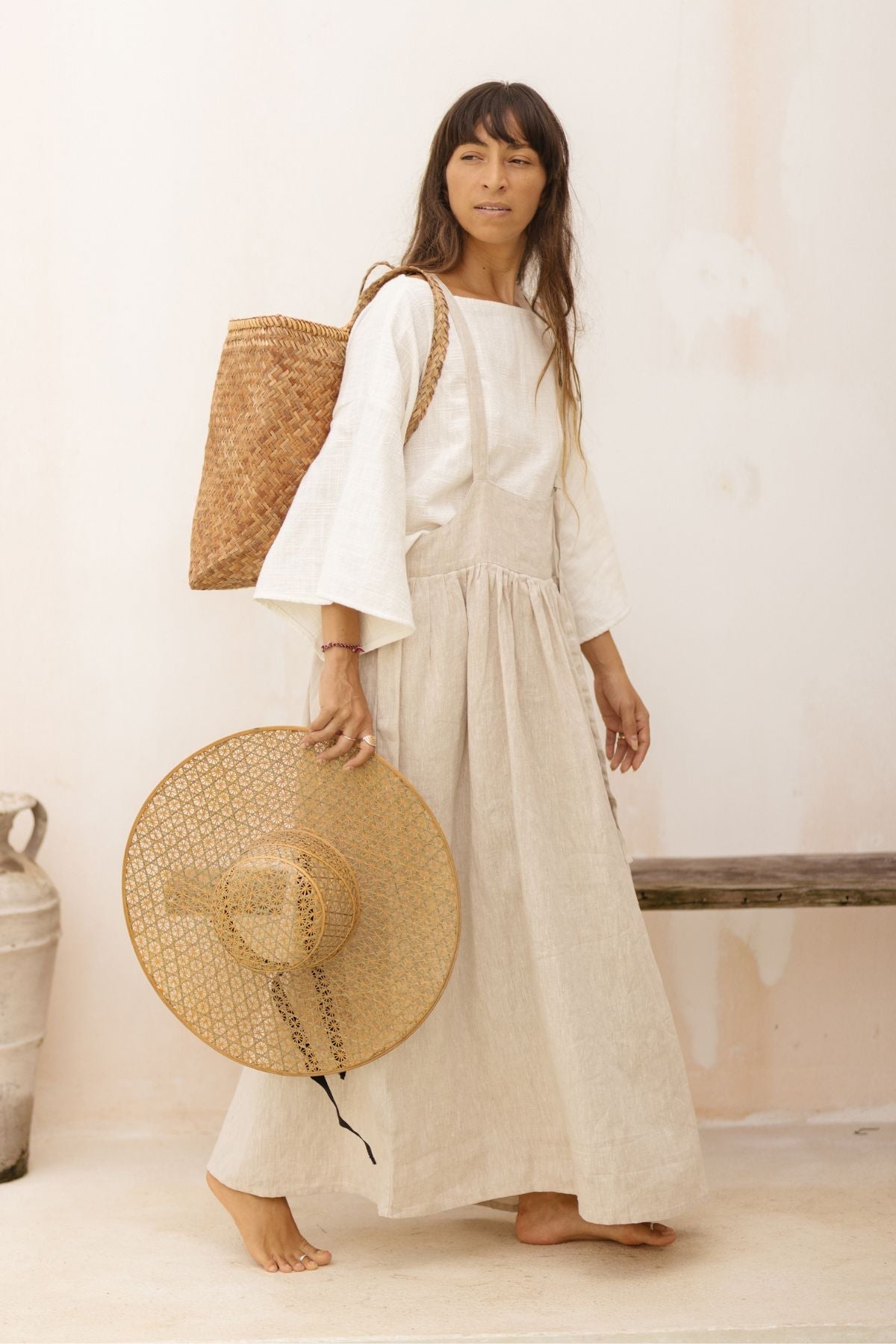 A woman stands barefoot on a concrete floor, holding a wide-brimmed woven hat and wearing the May May Overall Skirt (Made To Order) from Myrah Penaloza over a white top with flared sleeves. She has a large woven bag over one shoulder and is standing beside a rustic wooden bench.