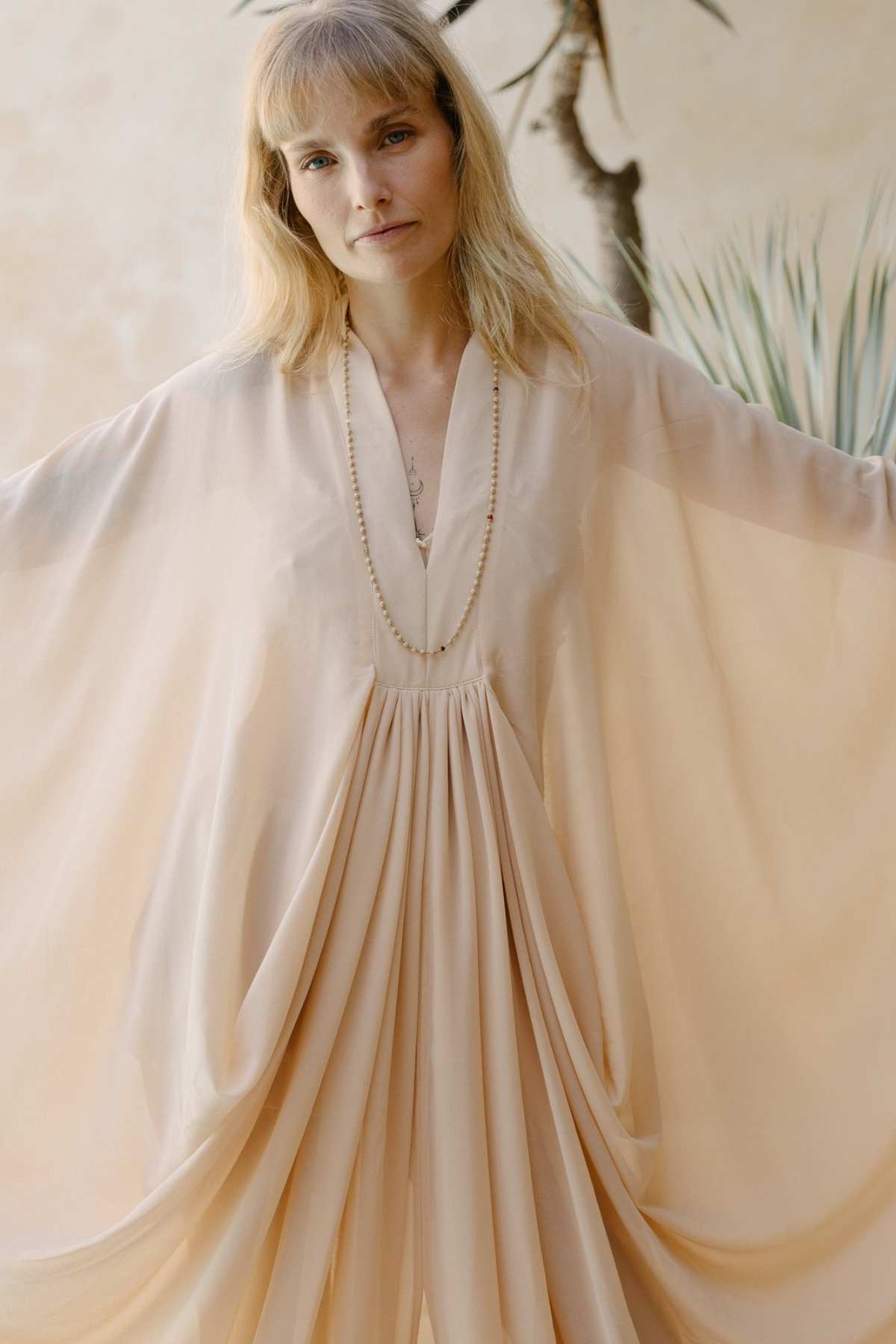 A person with long, blonde hair is wearing a luxurious Myrah Penaloza Leo Kaftan Chiffon Sheer in flowy, light beige chiffon silk with a deep V-neckline and draped sleeves. They are standing with arms slightly extended, displaying the fabric. The background features a blurred plant and a neutral-colored wall.