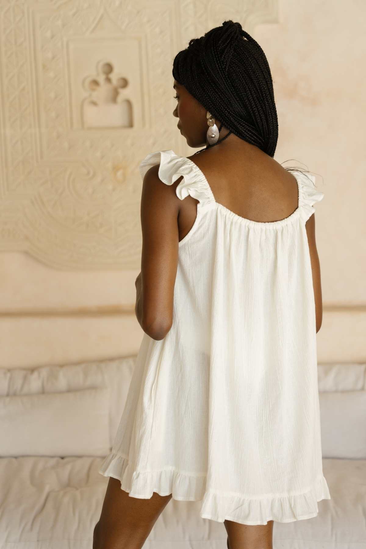 A woman with braided hair stands with her back to the camera, wearing a white Bumi Mini Slip Dress by Myrah Penaloza, adorned with ruffled edges. The background showcases light-colored, textured walls with intricate patterns.