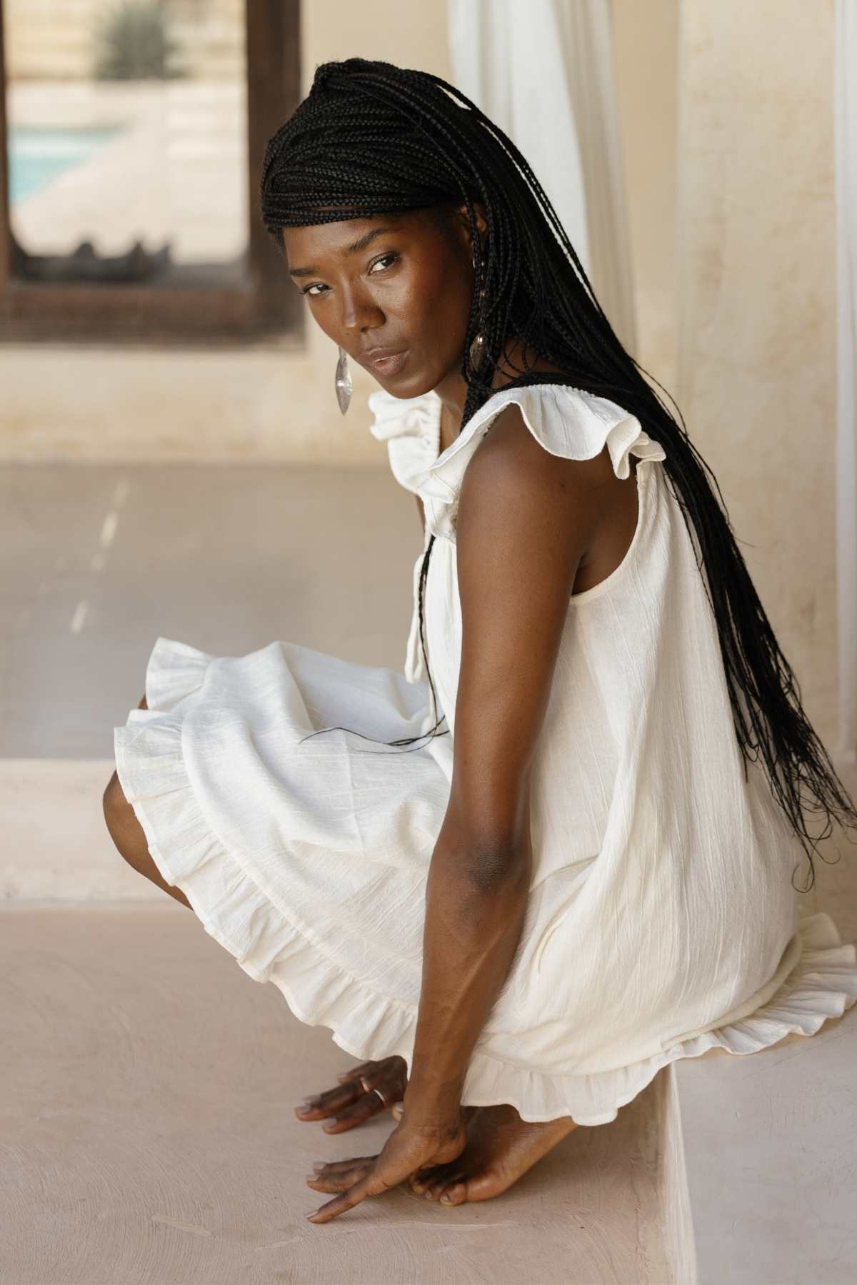 A woman with long braided hair, dressed in the Bumi Mini Slip Dress by Myrah Penaloza, is squatting on the floor indoors. She looks over her shoulder with a serene expression. The background is minimalistic with light-colored walls and a window.