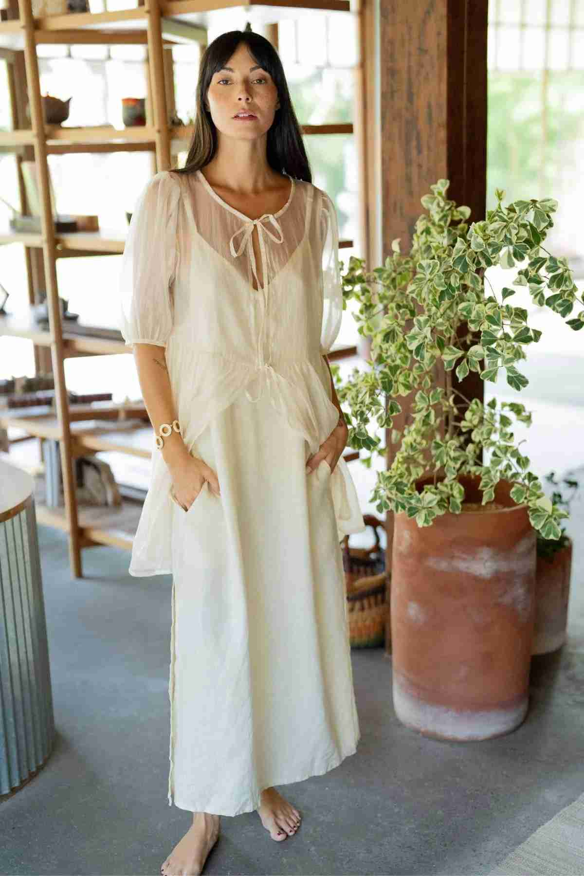 A woman with long dark hair stands barefoot on a concrete floor. She wears a Dynasty Sheer Silk Dress by Myrah Penaloza, gracefully tied at the neck, and has her hands in the pockets. Behind her is a large potted plant and a wooden shelving unit with various items, creating an elegant layering essential scene.