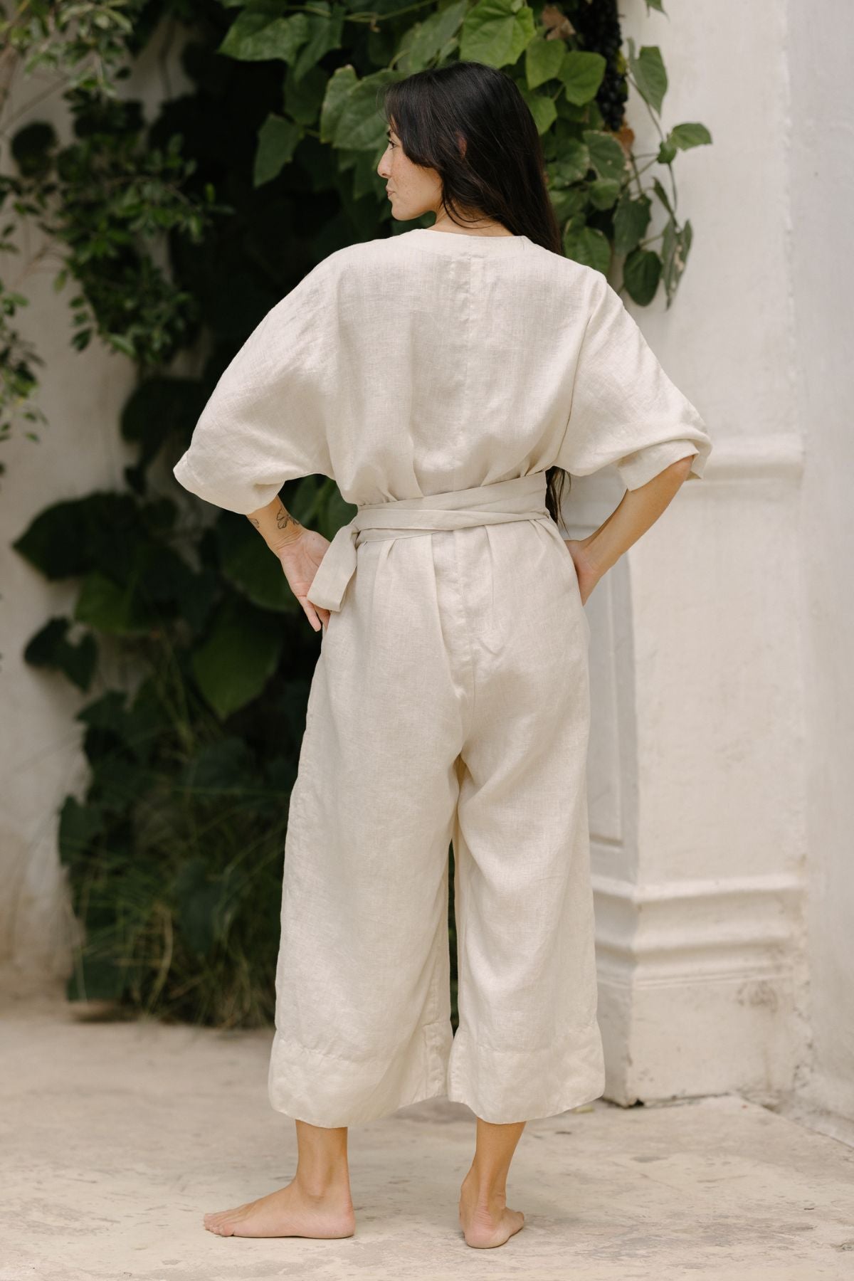 A person with long, dark hair stands barefoot, facing away from the camera. They are wearing the Rey Linen Playsuit by Myrah Penaloza, featuring short sleeves and a belt. The background showcases green plants and a white wall.