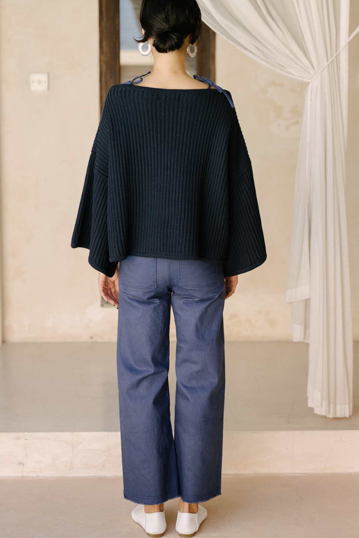 A person with short hair stands facing away from the camera, wearing an oversized Spirit Top Sweater Knit in dark blue with wide sleeves, crafted from luxurious cotton fabric. They are also dressed in light blue wide-leg pants and white socks. A partially visible white curtain serves as the background.