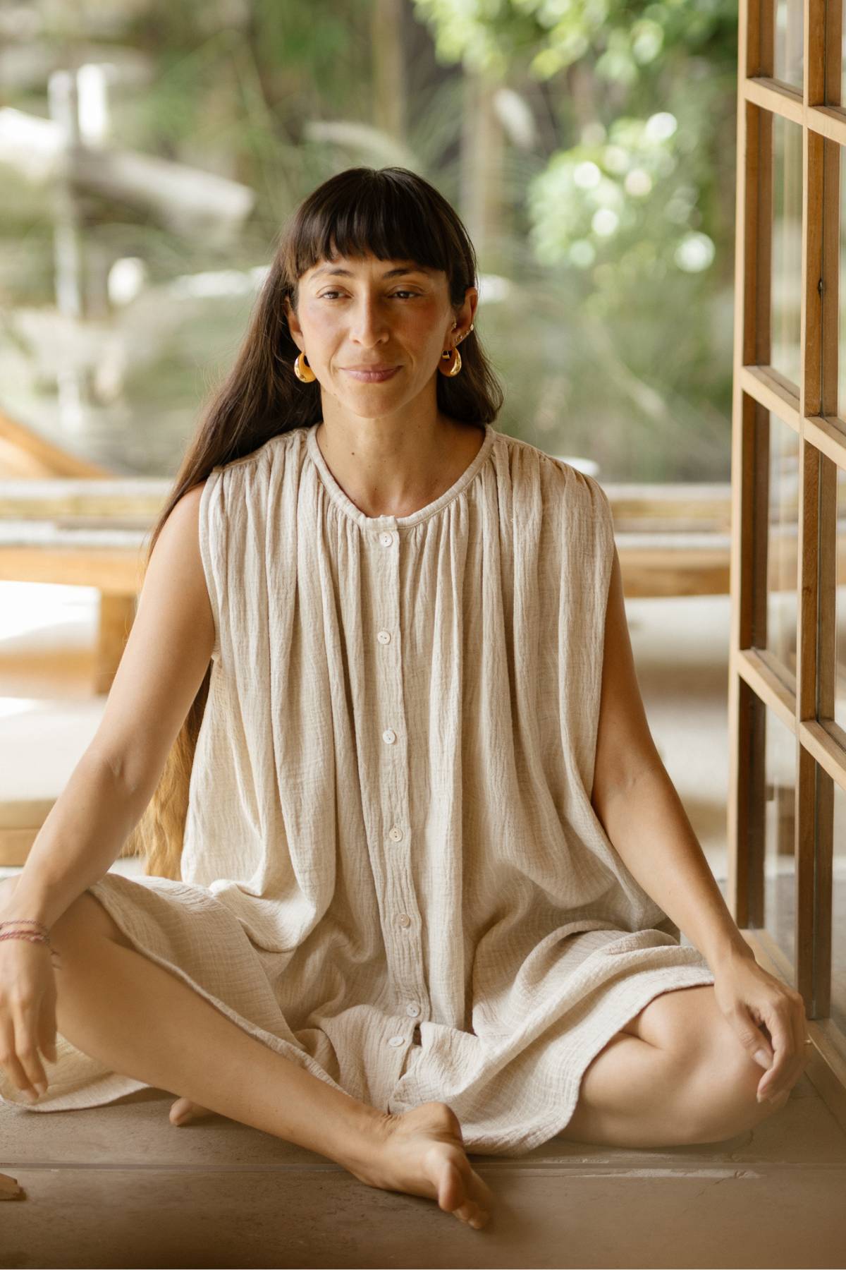 A woman with long hair and bangs sits cross-legged on the floor, wearing a loose, sleeveless Bonny Dress by Myrah Penaloza. Inside, she's positioned next to a wooden structure, with softly blurred greenery in the background. This cotton weave dress perfectly showcases her versatile style.