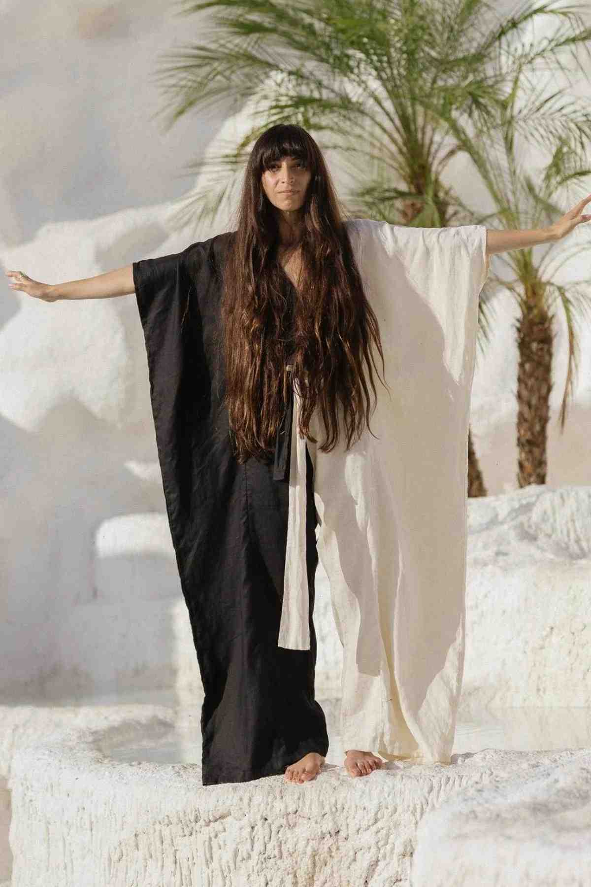 A woman with long dark hair stands with her arms outstretched, wearing the French Linen Yin Yang River Playsuit by Myrah Penaloza. This floor-length garment is split into two colors - black on the left side and white on the right side. She stands barefoot on a white rocky surface with a green palm tree in the background.