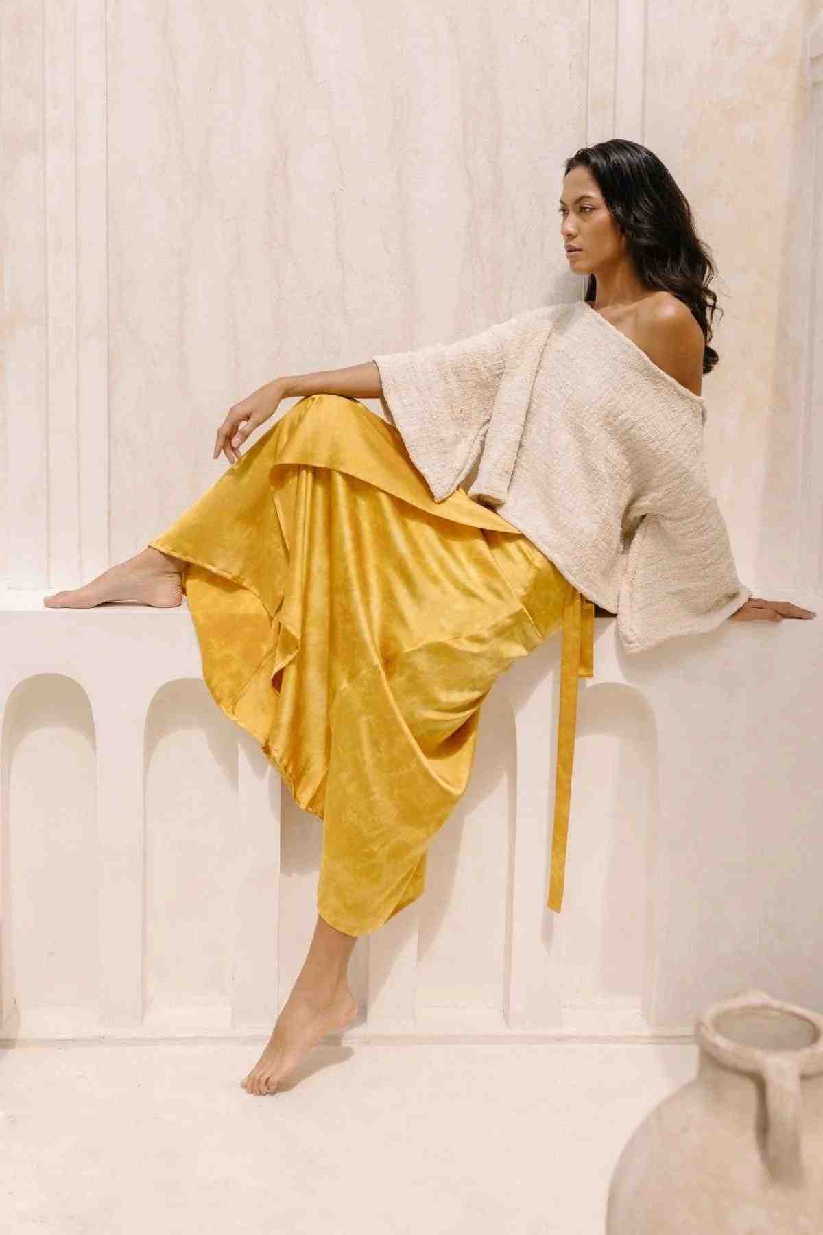 A woman sits gracefully on a white ledge against a light-colored background. She is wearing the Spirit Top & Shanti Wrap Skirt Set by Myrah Penaloza, which features a loose, off-the-shoulder cream sweater and flowing golden yellow pants made of hand-woven tummangal cotton. She looks to the side with one leg bent and the other extended, exuding a relaxed and elegant demeanor.