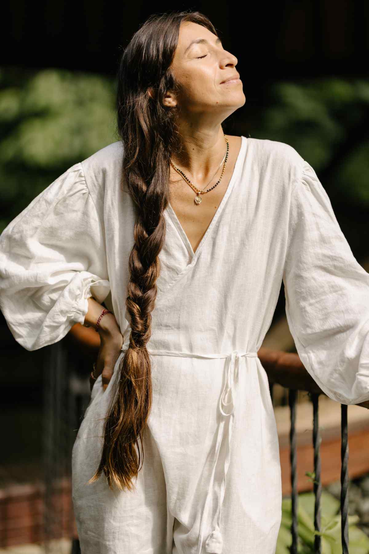 A woman with long, braided hair stands barefoot on a wooden deck amidst lush greenery. She is dressed in Myrah Penaloza's Linen Long Sleeve Dora Playsuit, showcasing its unique detailing and adjustable waistline for an elegant simplicity. She leans slightly against the railing, gazing contemplatively into the distance.