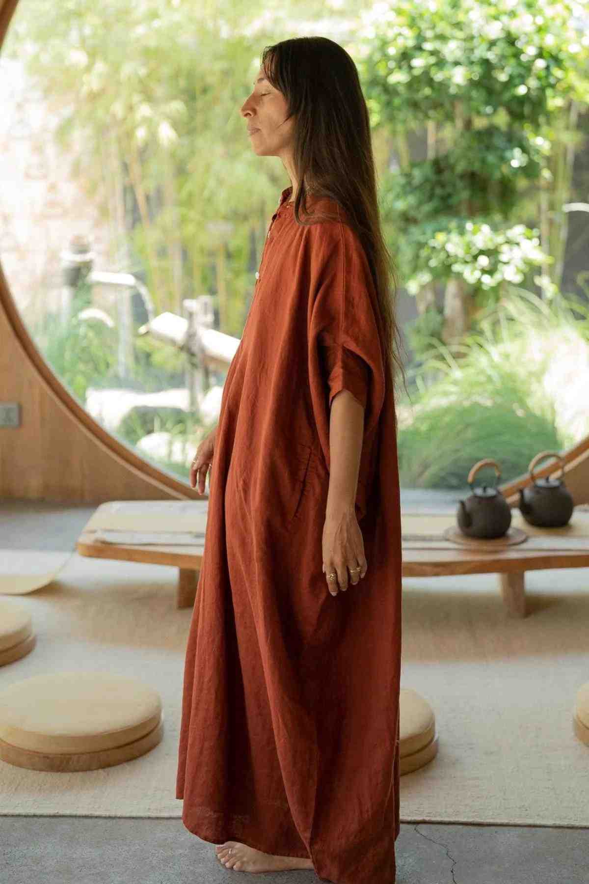 A person with long hair stands indoors, wearing a loose-fitting, rust-colored La Majia Kaftan by Myrah Penaloza. They are barefoot and appear relaxed. Behind them, a round window offers a view of a lush garden with various green plants and a small wooden table with two teapots, evoking the spirit of Frida Kahlo's serene retreats.