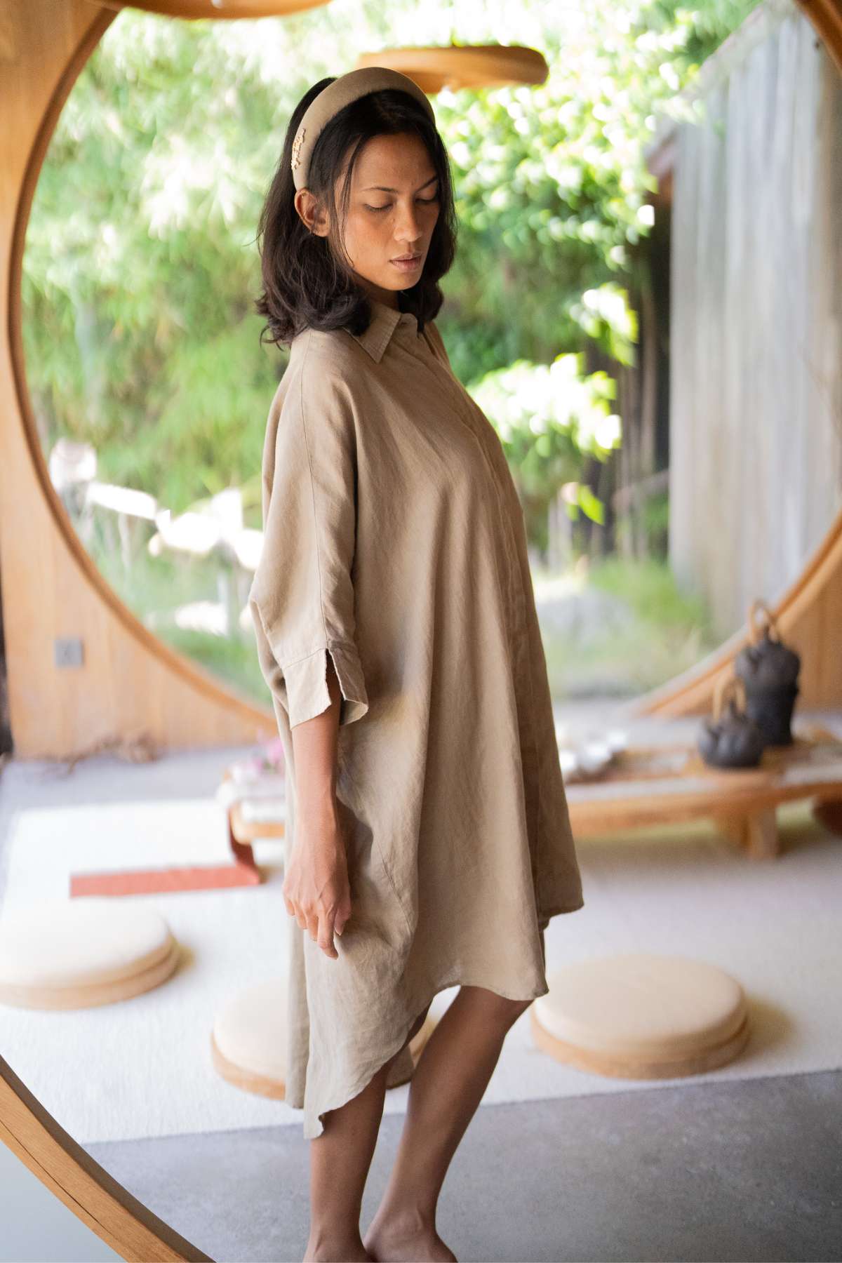 A woman in the Linen Suka Button Down Long Top - BF by Myrah Penaloza stands inside a circular doorway, enveloped by a serene, nature-inspired setting adorned with soft lighting and wooden accents. Her dark hair is complemented by a headband, showcasing versatile styling options while highlighting the elegance of sustainable fabric in this tranquil scene.