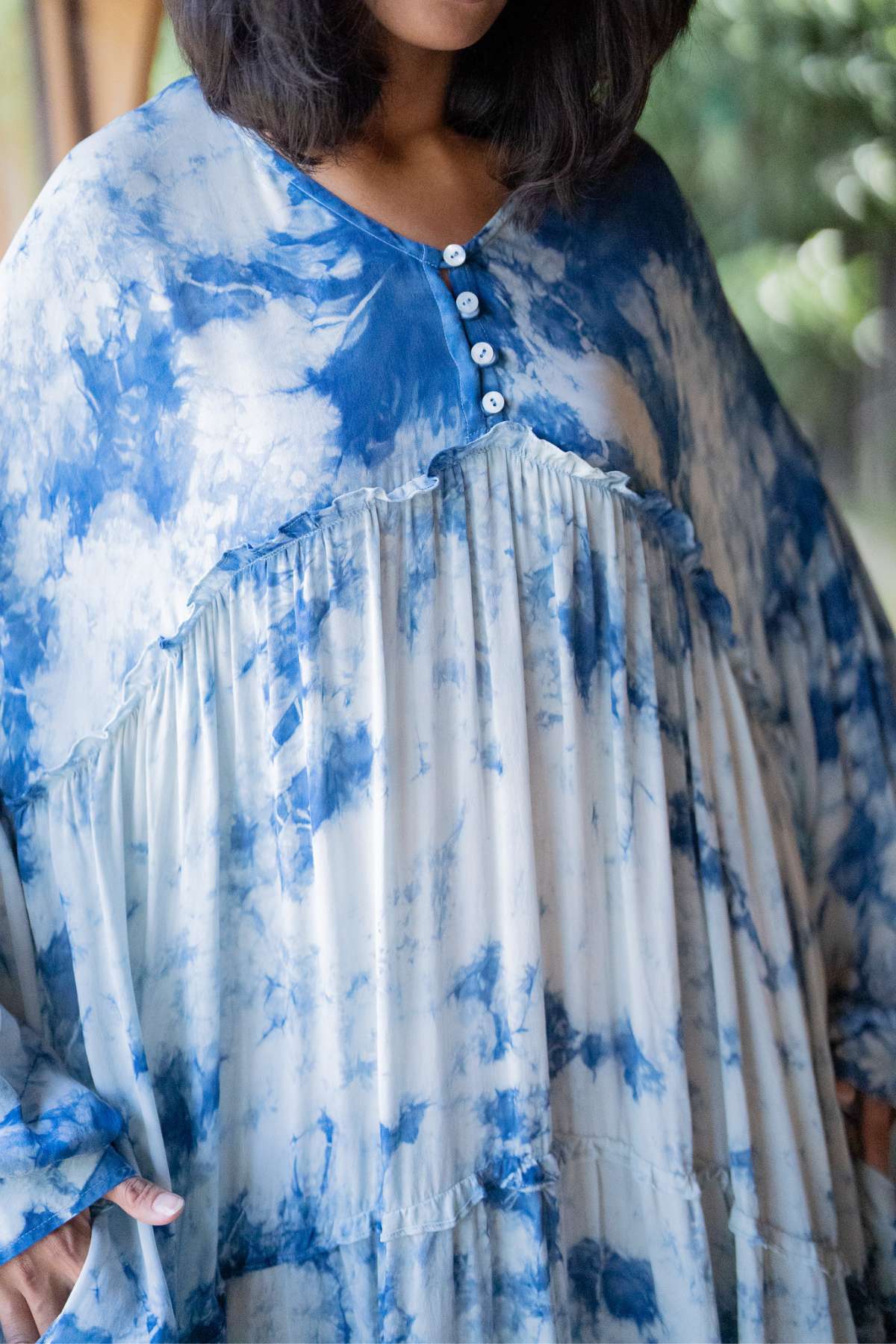 A person wearing the Indigo Eva Gown from Myrah Penaloza, crafted from 100% silk. The dress is indigo-dyed, showcasing button detailing at the neckline, an oversized, loose fit, and a ruffled design. The background is slightly blurred with hints of greenery.