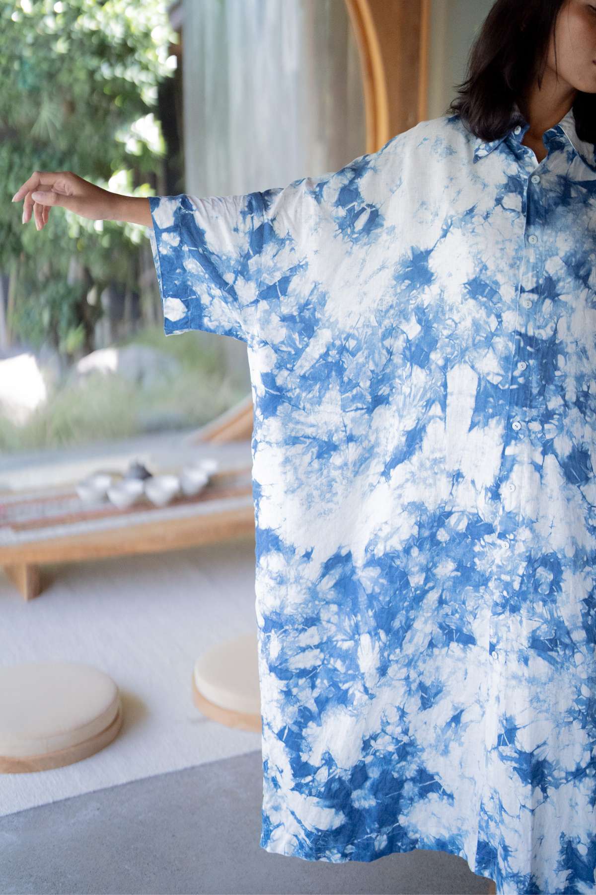 Someone wearing The Boyfriend Dress - Hand Tye Dyed Indigo by Myrah Penaloza, with their arms slightly spread out. The backdrop includes a tranquil indoor setting with natural tones, round cushions, and greenery visible through a window.