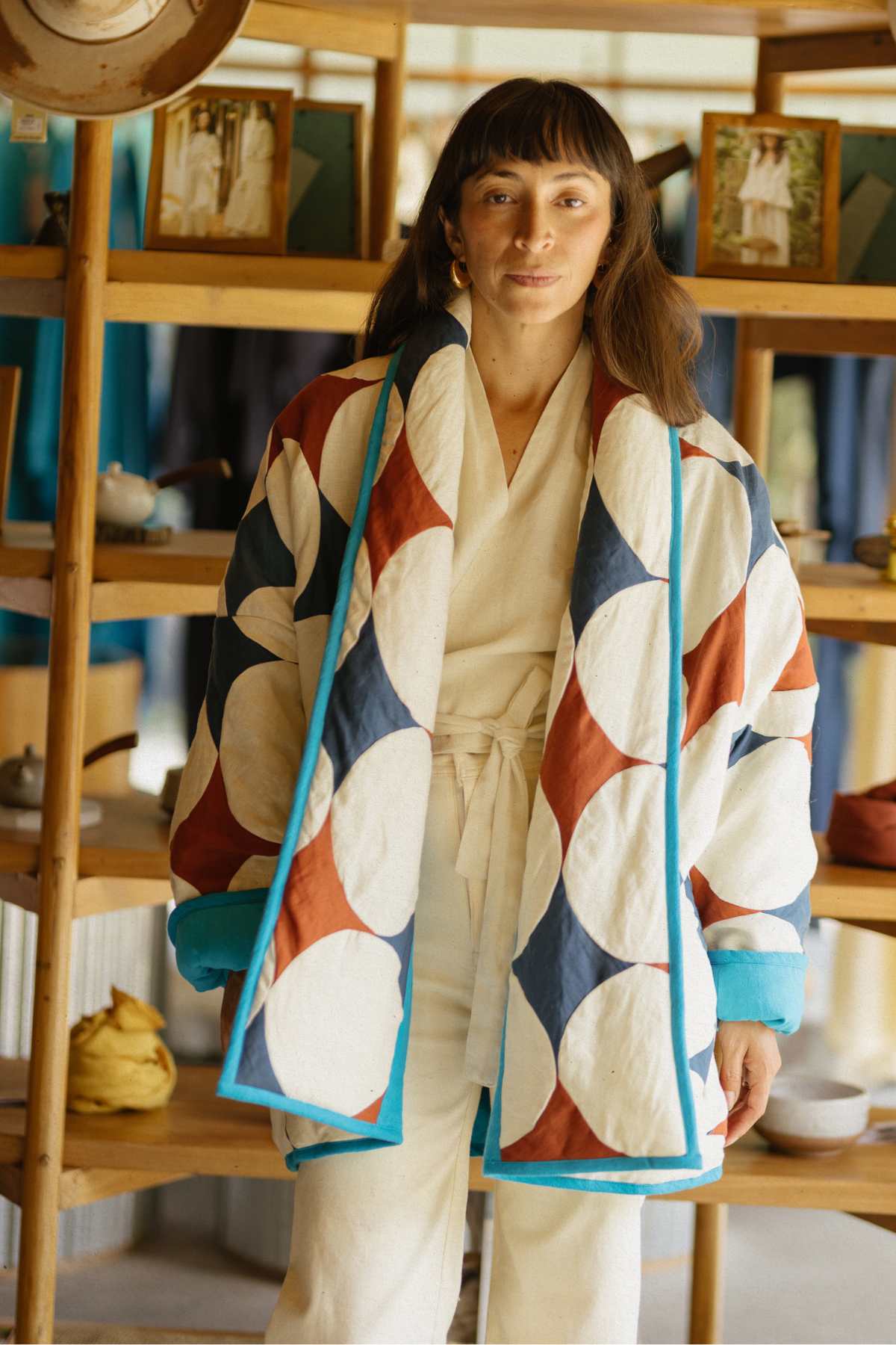 A woman with long brown hair and bangs stands in a store, wearing the Javi Quilted Jacket from Myrah Penaloza, decorated with large geometric patterns in blue, brown, and white over a white outfit. Behind her are wooden shelves displaying various items, including clothing and decor.