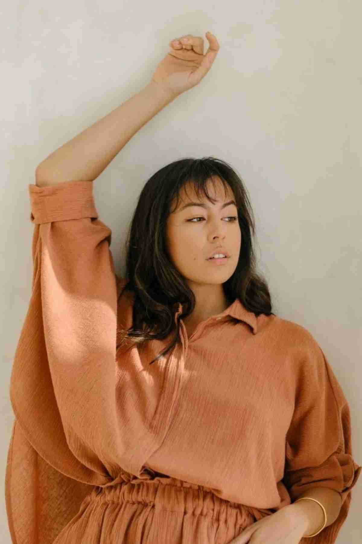 A woman in a Suka Button Down Top by Myrah Penaloza, handmade in Bali, leans thoughtfully against a light wall. Her right arm is raised above her head, and her dark hair falls to her shoulders. The soft, warm lighting casts gentle shadows, highlighting the top's sustainable fashion and plant-dyed hues.