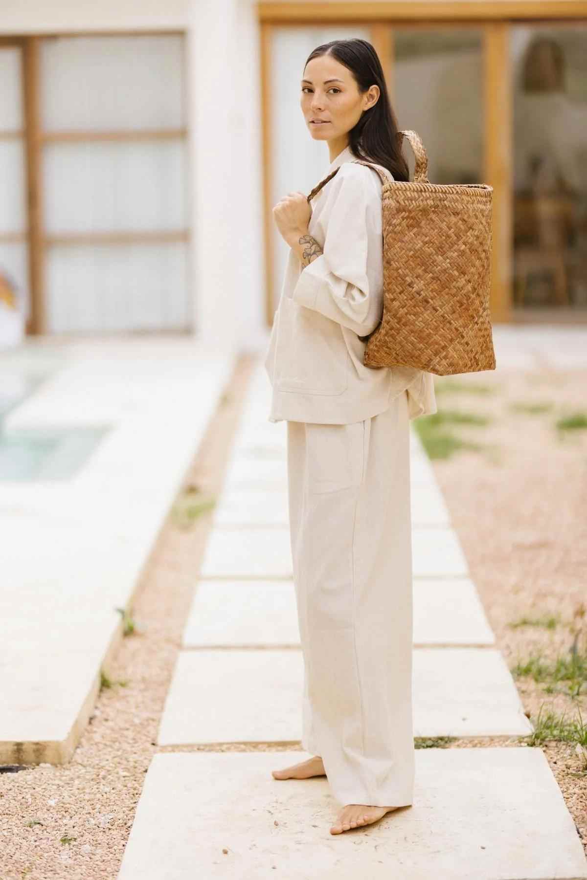 A woman stands outdoors on a stone pathway wearing the loose-fitting, natural cotton-linen Grace Jacket & Mona Pant Set by Myrah Penaloza. She carries a large woven basket backpack and looks at the camera, with the backdrop featuring large windows and a glass door of a modern building.