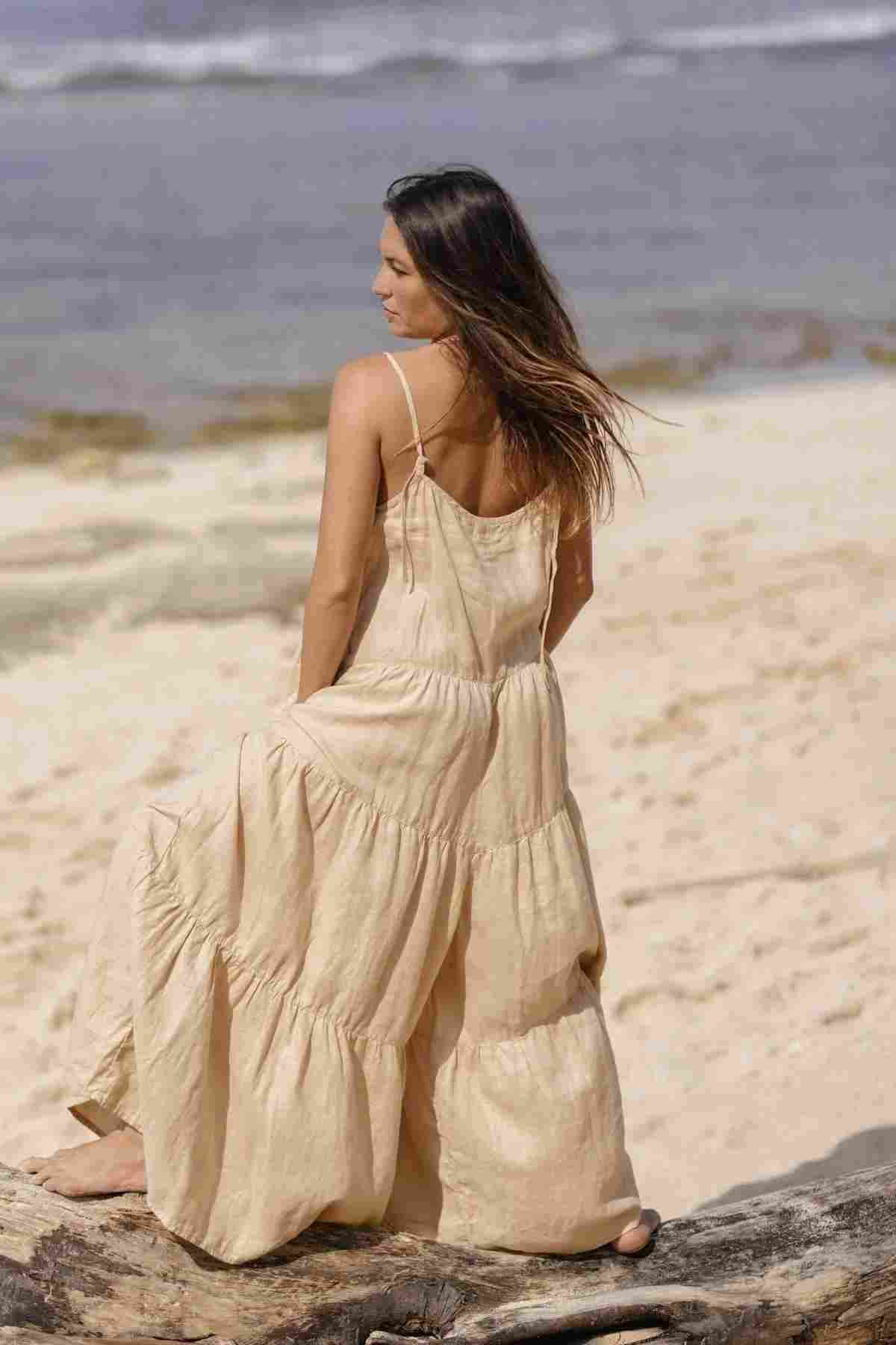 A woman in a custom-made La Mexicana Playsuit from Myrah Penaloza, crafted from 100% linen, stands on a rocky ledge at the beach. Her long hair gently blows in the wind, creating an elegant silhouette against the serene backdrop of sand and waves.