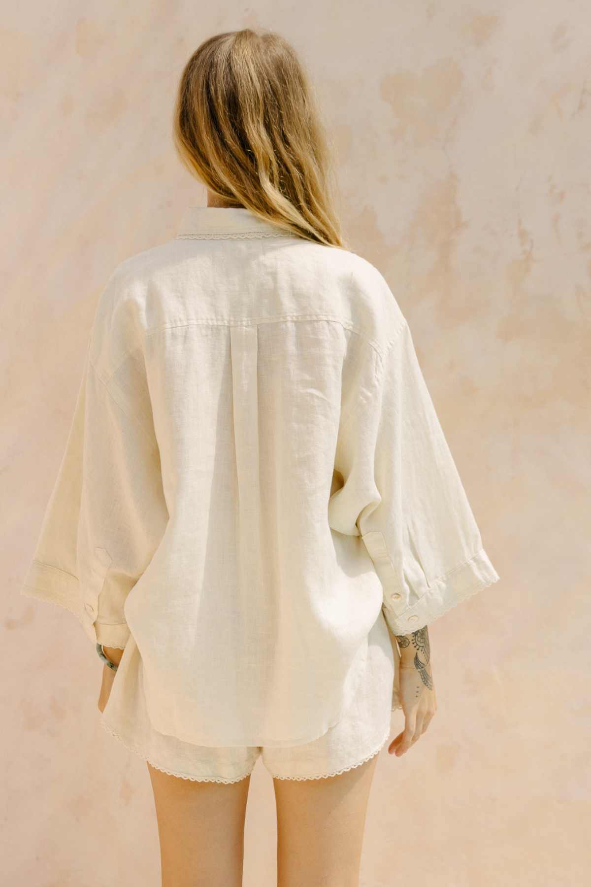 A person with long hair stands with their back to the camera, wearing an off-white, loose-fitting Nidra Button Down Set MoonLight Linen from Myrah Penaloza, made from organic cotton. The shorts perfectly complement the ensemble against a soft, light-textured background with warm tones.