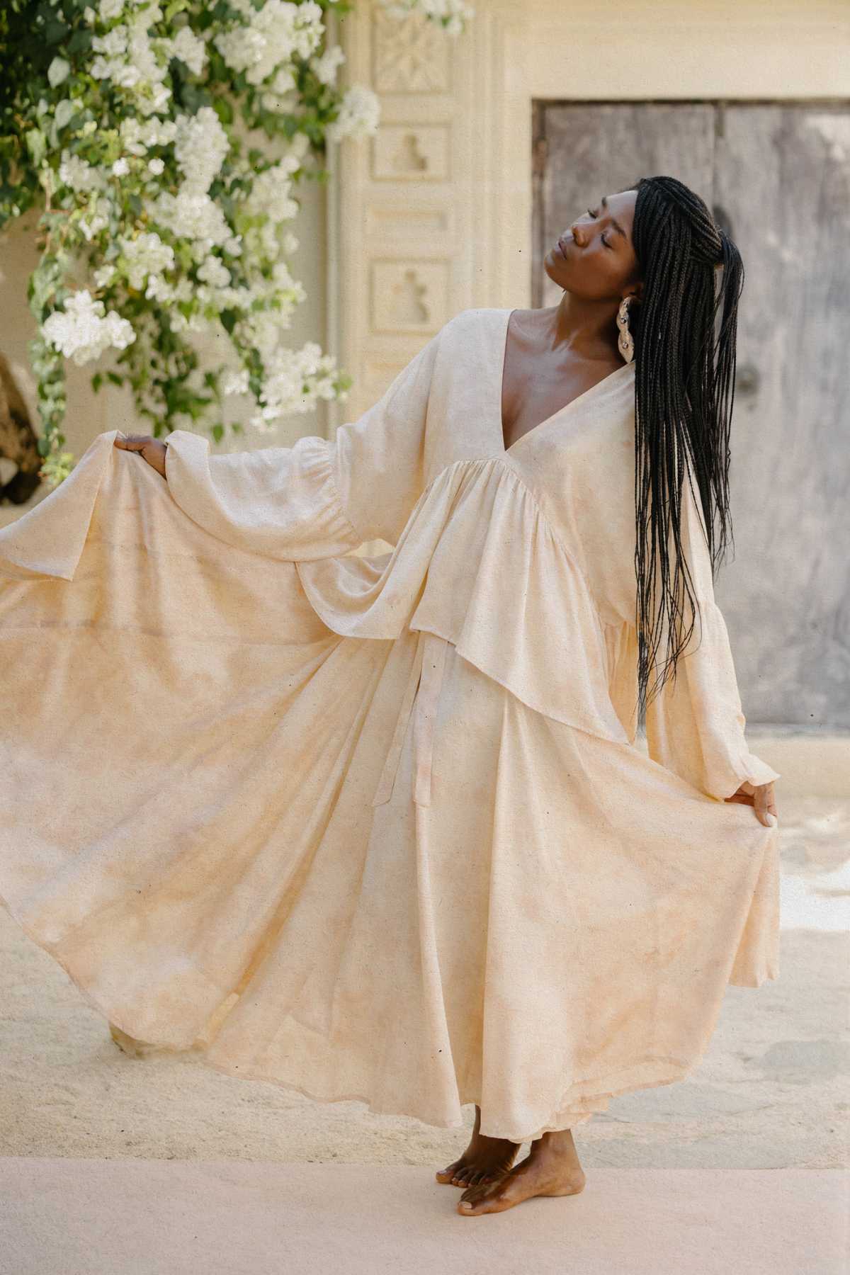 A woman gracefully poses barefoot on a sandy surface, modeling the elegant Eva Top & Orisa Dance Skirt Set by Myrah Penaloza. Her braided hair falls to one side as she closes her eyes, surrounded by lush greenery and a rustic wooden door in the background.