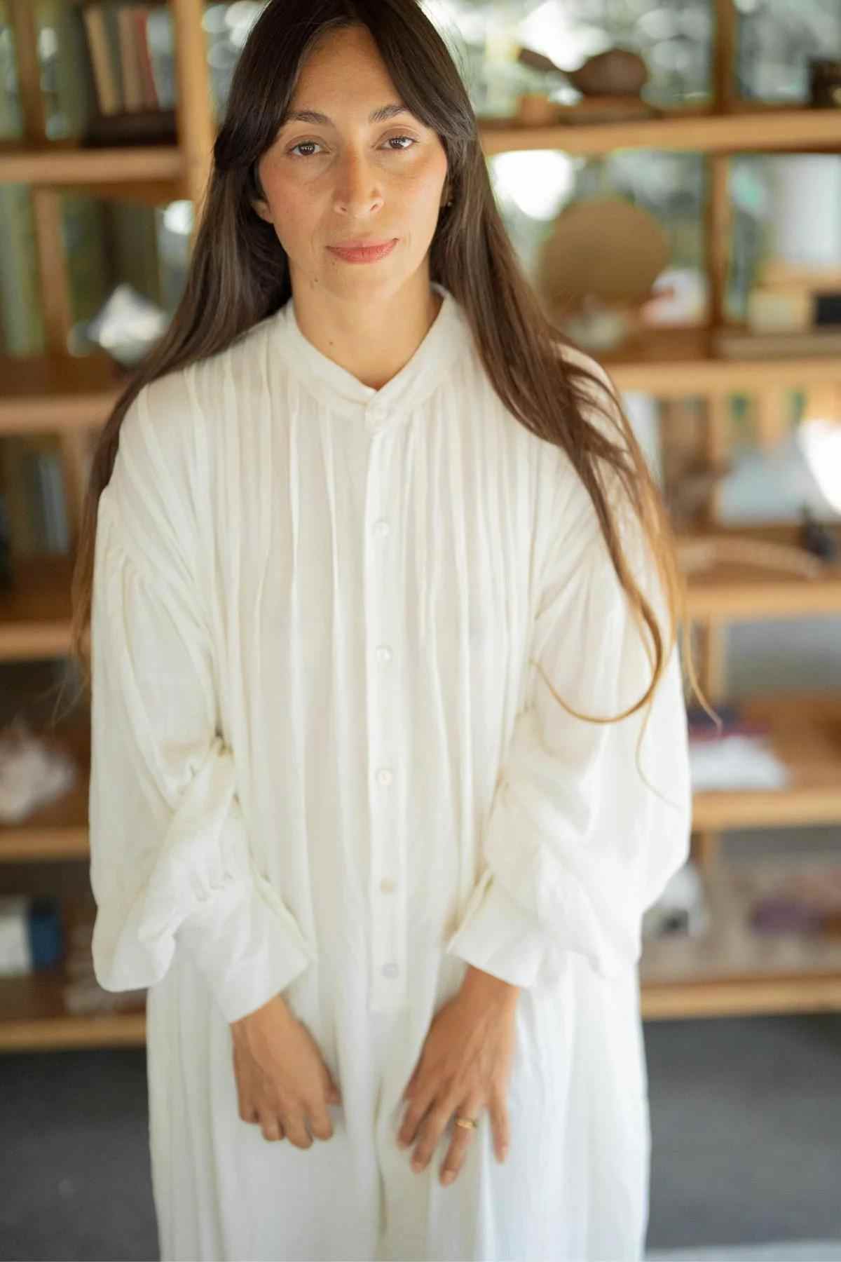 A woman with long brown hair stands in front of a wooden shelf unit. She is wearing the newly released Kundalini Gown Playsuit Edition (Pre-Order) by Myrah Penaloza, which features elegantly gathered sleeves. Facing the camera, she is sharply in focus against a softly blurred background, making her the center of attention in the image.