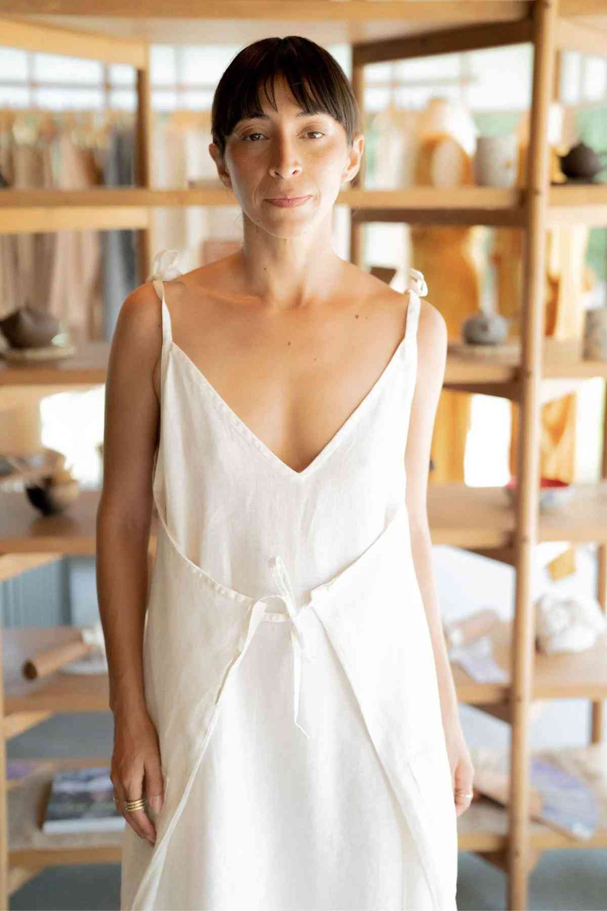 A woman wearing a white, sleeveless Radiant Gown - Made To Order by Myrah Penaloza stands in a room with wooden shelves in the background, filled with various items like pottery and fabrics. With her short dark hair and bangs, she exudes a minimalist style as she looks directly at the camera, embodying the elegance of our healing linen collection.