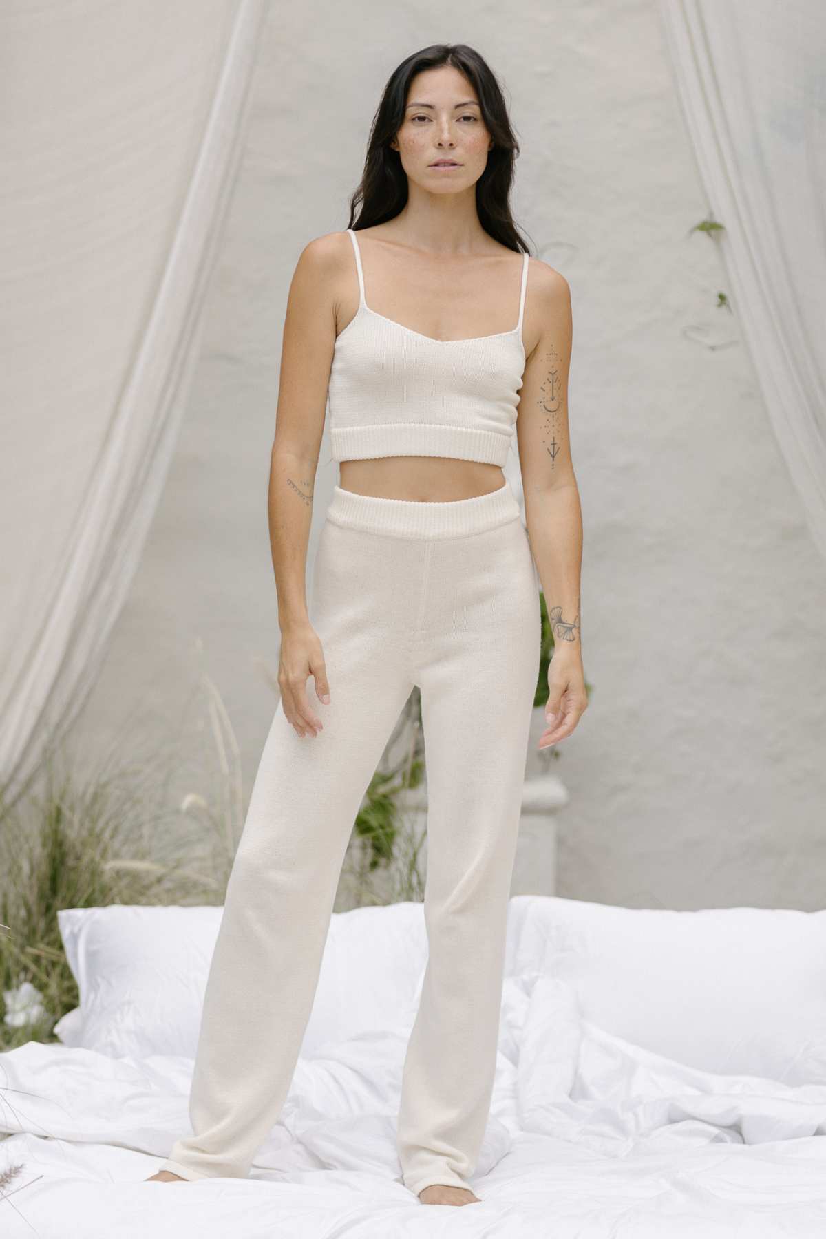 A person dressed in a cream-colored, sleeveless crop top paired with matching high-waisted pants from the Honey Knit Sets by Myrah Penaloza poses stylishly on a white bed with draped fabric in the background. This chic outfit is made using sustainable cotton, and sparse greenery adds to the setting.