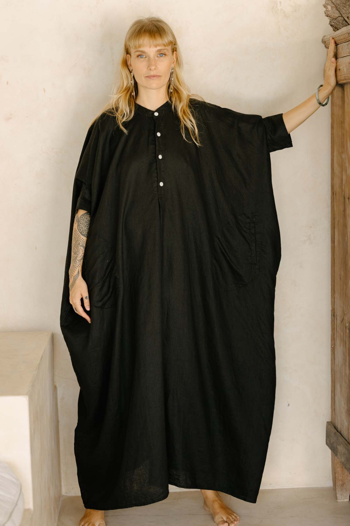 A person with long blond hair stands barefoot on a light floor, wearing the La Majia Kaftan by Myrah Penaloza. Their left arm rests on a wooden frame, and their right arm is relaxed by their side. The background features light-colored walls and a rustic wooden element.