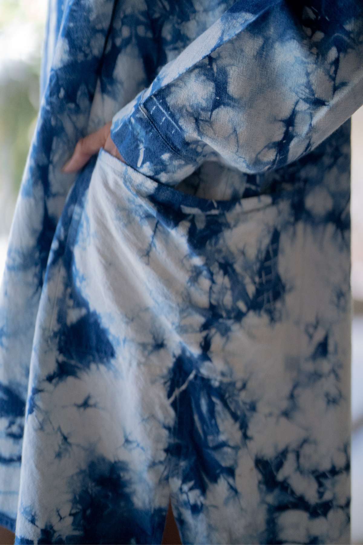 A close-up captures someone wearing the Tender Jacket by Myrah Penaloza, hand-dyed in indigo. One hand rests in its pocket, revealing a marbled tie-dye pattern against a softly blurred background. This remarkable piece perfectly blends artistry with style.