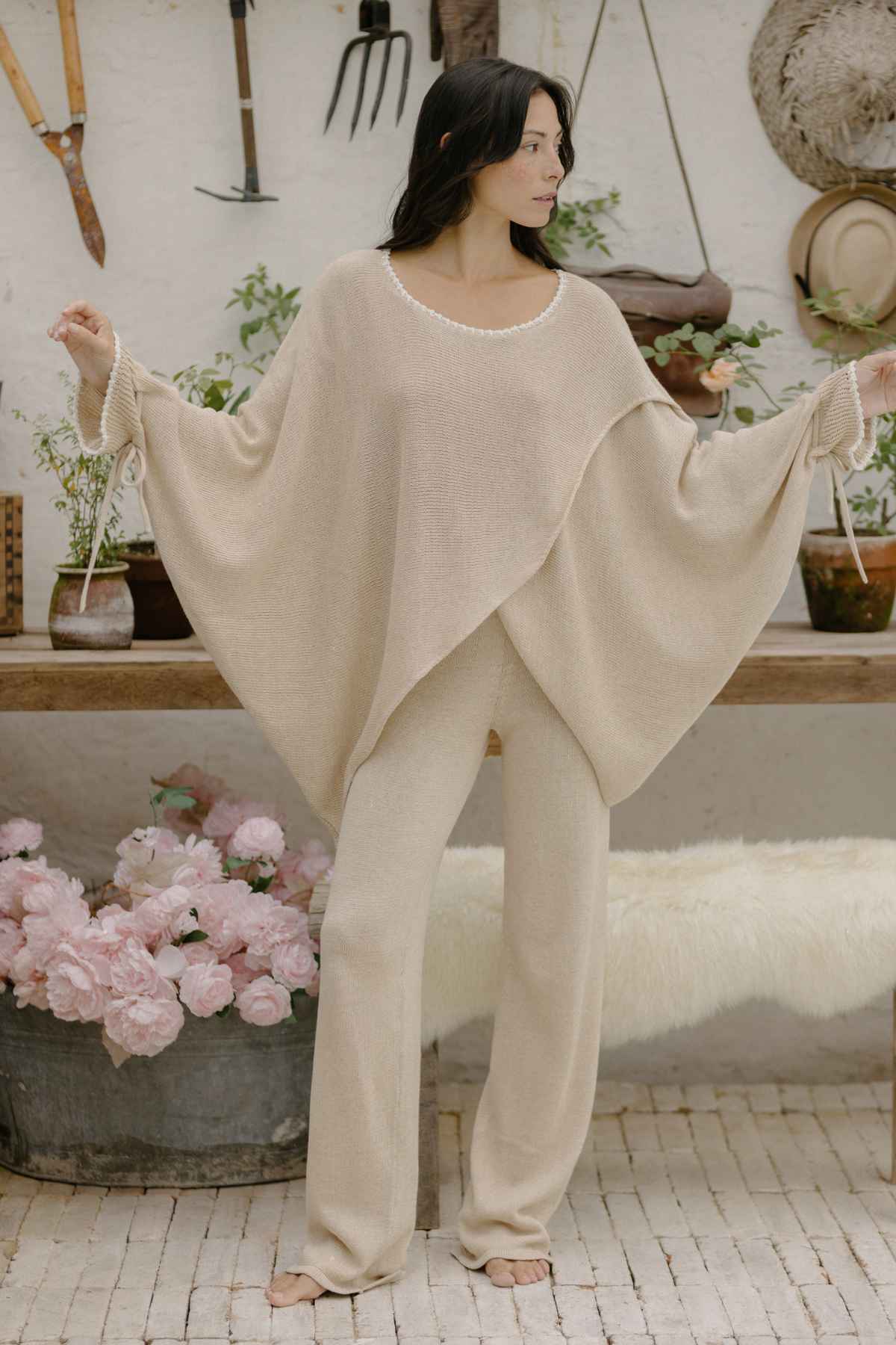 A person with long dark hair stands indoors wearing the loose, beige Honey Knit Sweater from Myrah Penaloza and matching pants. They are holding the sweater’s extended sleeves. The background features gardening tools, a hat, plants on a shelf, and a basket of pink flowers.