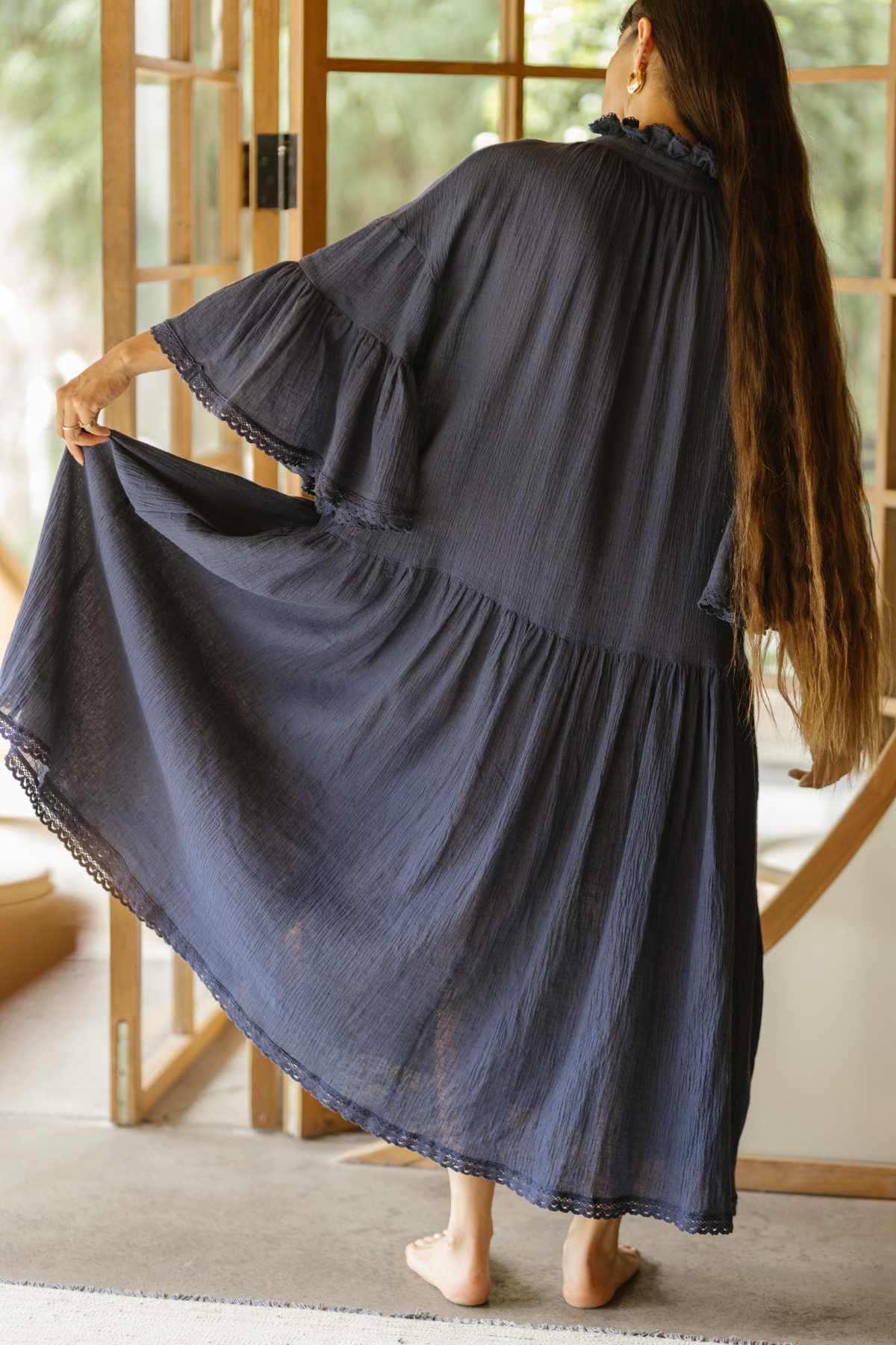 A person with long hair stands barefoot indoors, elegantly dressed in a flowing Kyoto Duster by Myrah Penaloza, which resembles ceremonial attire. They gently hold the edges of the dark blue dress, highlighting its intricate pleats. In the background, wooden doors and a blurred green scenery are visible.