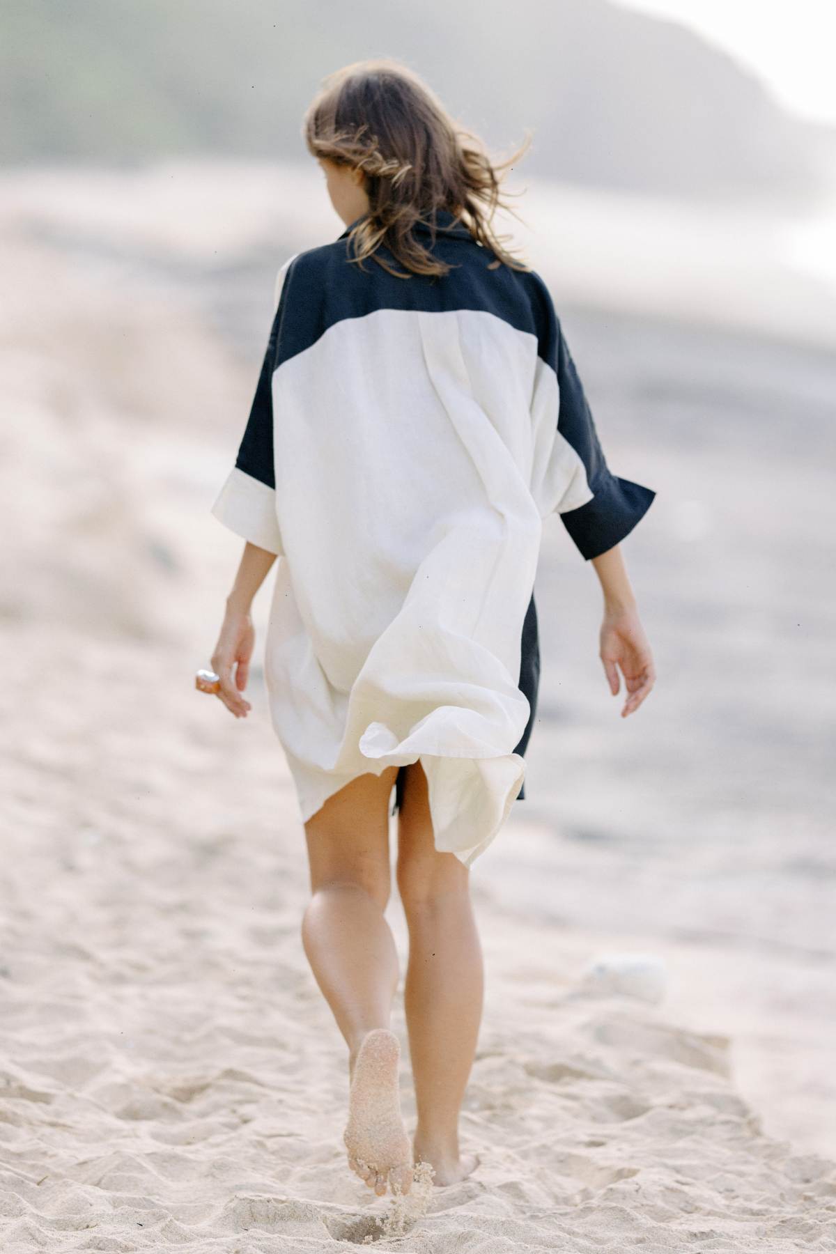 A person with shoulder-length hair walks barefoot on a sandy beach. They are wearing the Linen Suka Button Down Long Top by Myrah Penaloza, a loose, black and white dress made from sustainable fabric. The shoreline and some greenery are visible in the background, creating a serene beach scene.