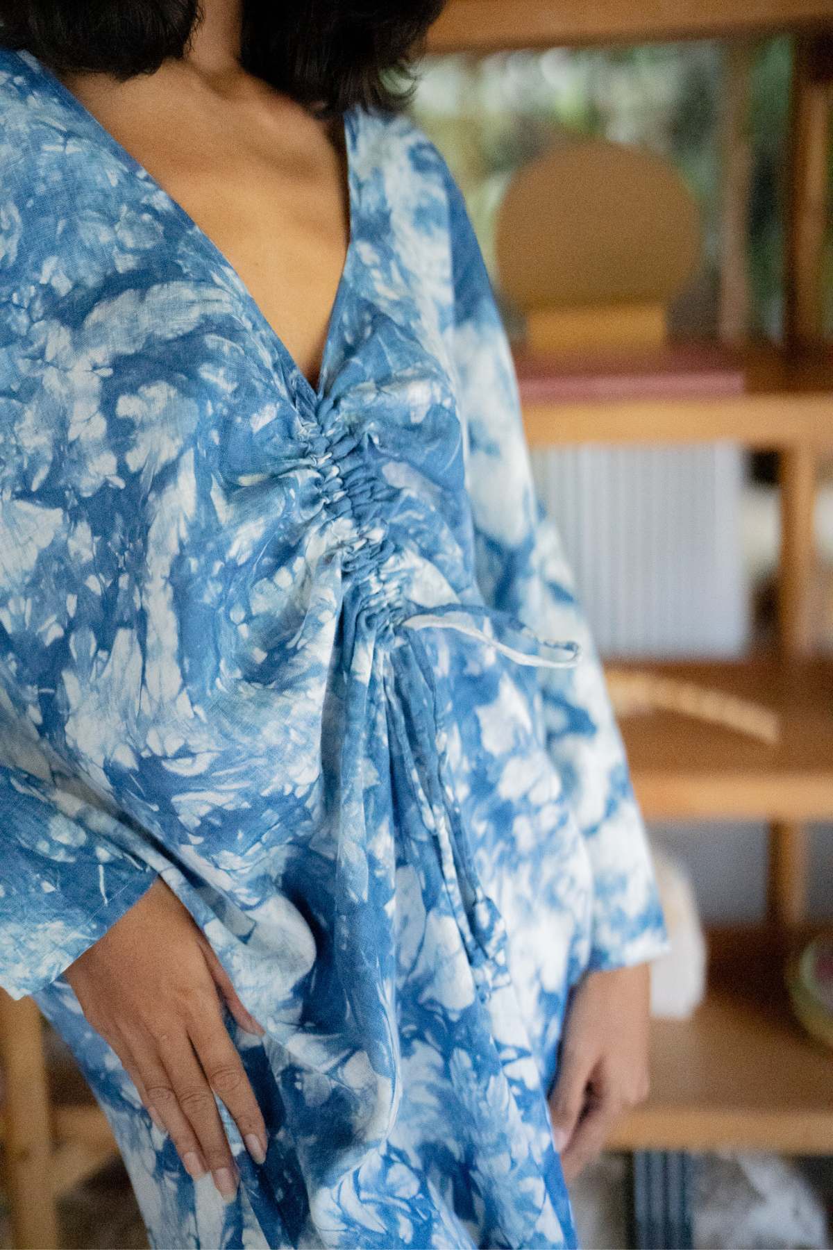 An individual is seen indoors wearing the one-of-a-kind Indigo Virgo Rising Kaftan by Myrah Penaloza, showcasing its unique blue and white tie-dye design with gathered fabric details. The dress's texture and patterns are in sharp focus, while the background features blurred shelves adorned with decor items.