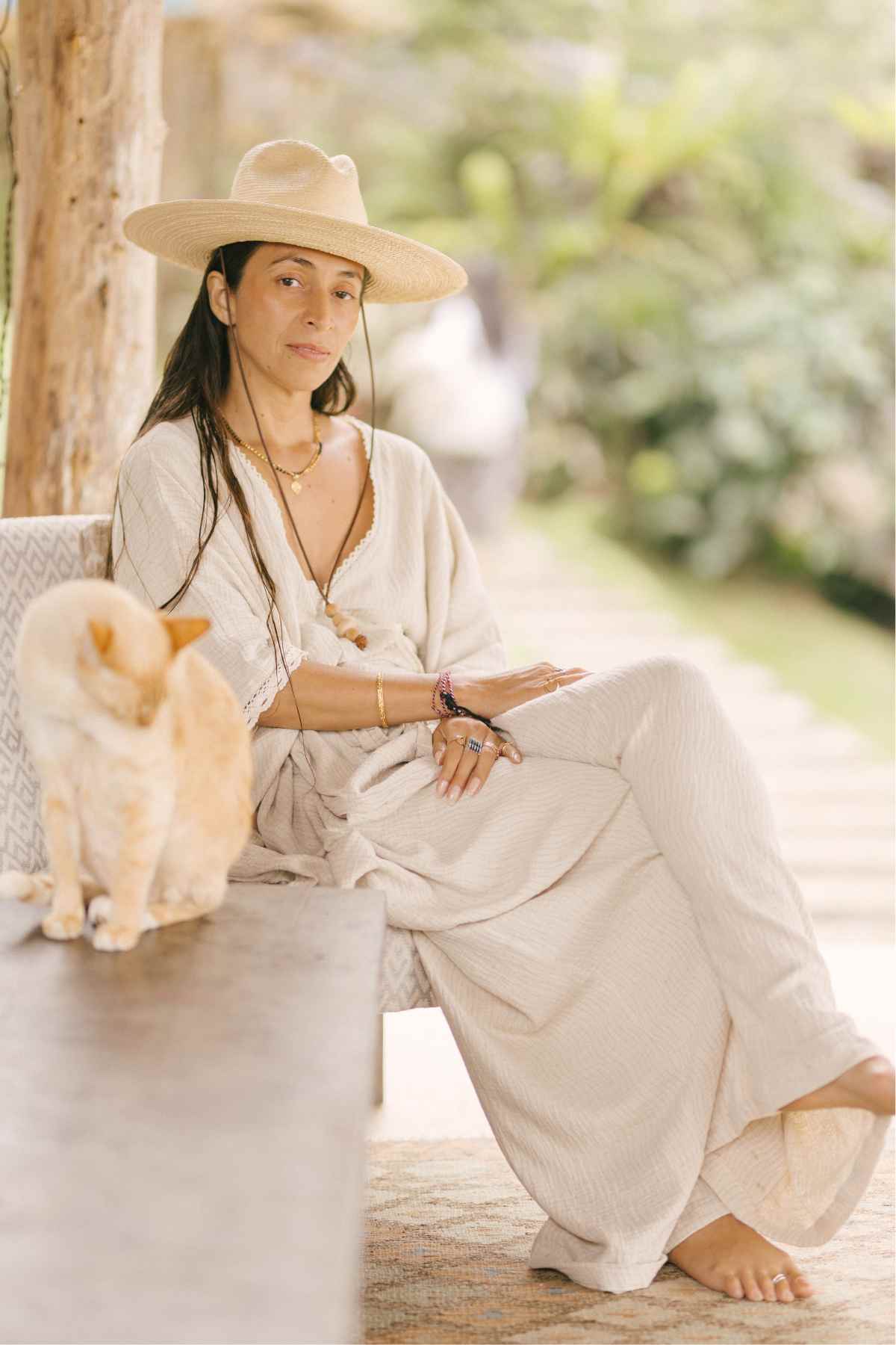 A woman in a Limited Edition Hand Embroidered River Playsuit by Myrah Penaloza and a wide-brimmed hat sits on a bench outdoors, surrounded by lush greenery. She is adorned with hand-embroidered necklaces and bracelets, her long hair braided. Beside her, a ginger cat lounges contentedly. The scene evokes the elegance of a limited edition moment in time.