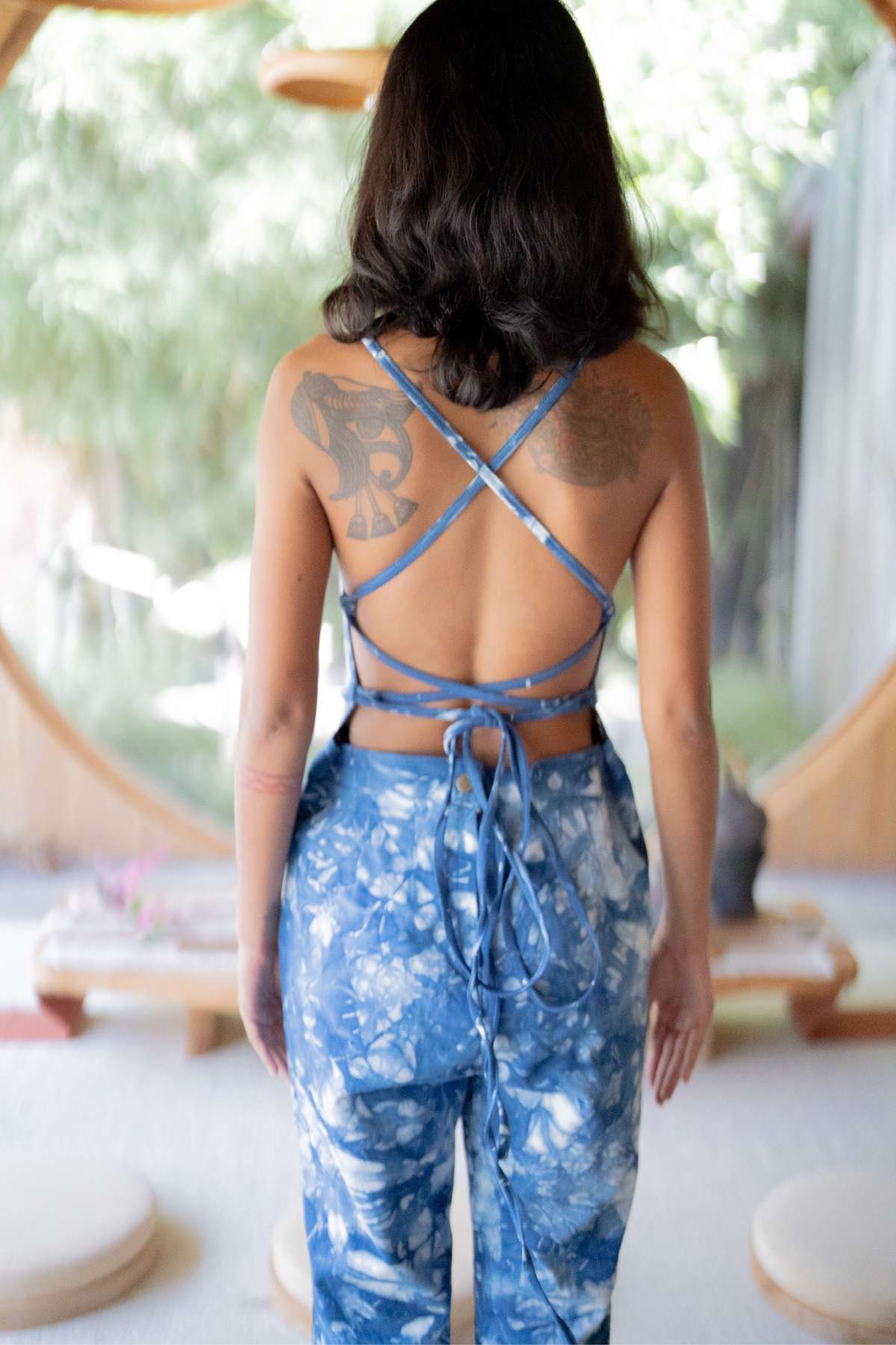 A person with tattoos is seen from behind, wearing the Indigo Dreams Camila Playsuit by Myrah Penaloza, a hand tie-dyed indigo backless playsuit with crisscross straps. The setting includes a round window and wooden furniture, making it an enchanting backdrop for this ideal date night ensemble.