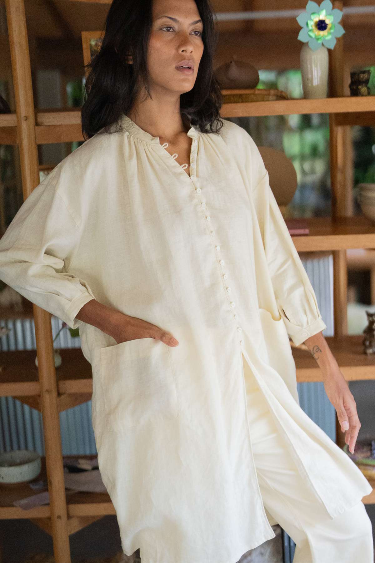 A person wearing a cream-colored, loose-fitting Zazen Linen Top by Myrah Penaloza stands in a room. The top exudes effortless elegance with its long sleeves and buttons. Pockets add practicality, while wooden shelves display pottery and decor items in the background.