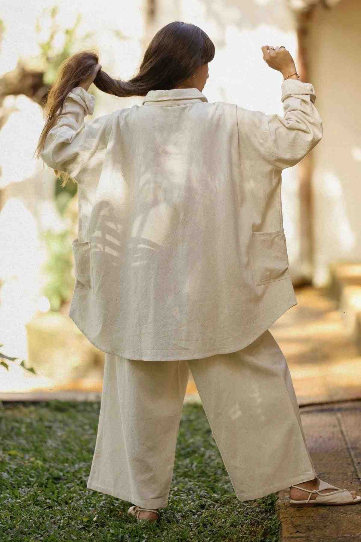 A person stands outdoors facing away, holding their hair up with both hands. They are clad in Myrah Penaloza's oversized light beige Tender Jacket (100% Linen) featuring large pockets on the sides, paired with matching wide-legged pants. The background is blurred, giving a hint of greenery and sunlight.