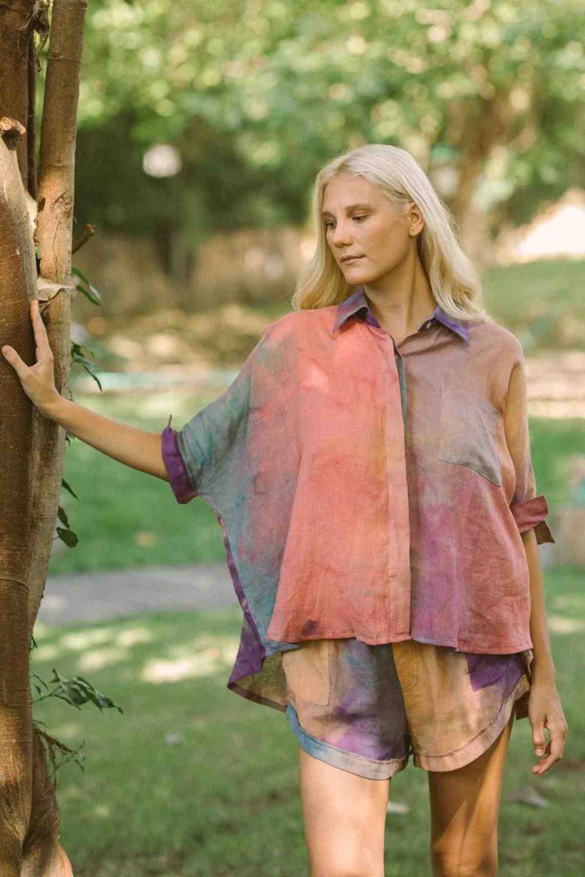 A person with long blonde hair is outdoors in a grassy area, wearing the vibrant and loose-fitting Suka Button Down Top from the Rainbeau Collection by Myrah Penaloza and handmade shorts. They are leaning against a tree with one hand, looking off to the side. The scene is sunny and tranquil.