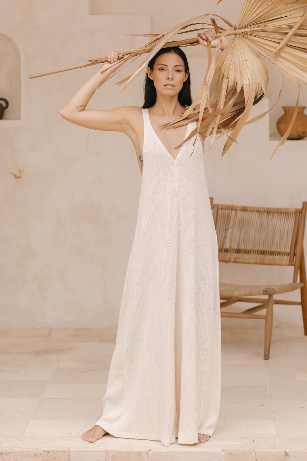 A woman stands on a light-colored tiled floor, wearing the Kundalini Playsuit—a limited edition cotton knit piece from Myrah Penaloza that embodies versatile elegance. She holds large dried palm leaves above her head against a neutral beige background with a wooden chair in the corner.