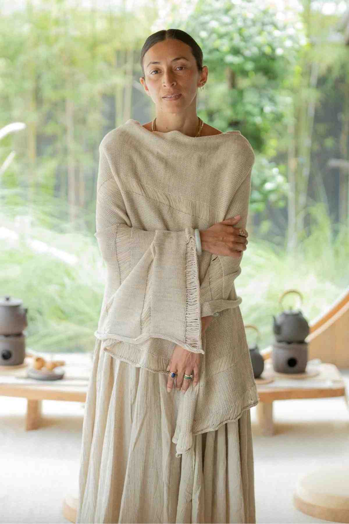 A person with dark hair pulled back, wearing the Frida Poncho from Myrah Penaloza—a light beige shawl and skirt ensemble that epitomizes effortless chic—stands with arms crossed. The background showcases a light-filled space adorned with wooden elements and greenery, including bamboo and plants, creating a serene and natural atmosphere.