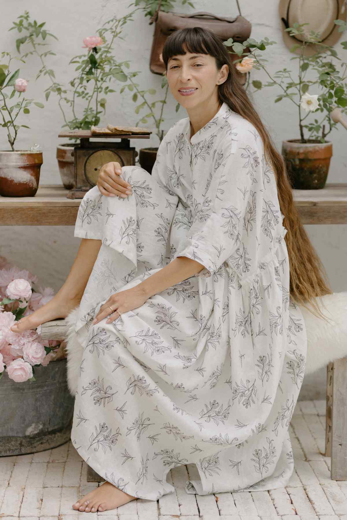 A woman with long hair sits barefoot on a chair in a rustic garden setting. She is wearing a light, Bona Dress from the Cha Dao Tea Collection by Myrah Penaloza and has a relaxed smile on her face. Around her, there are potted plants and flowers, creating a calm and natural atmosphere.