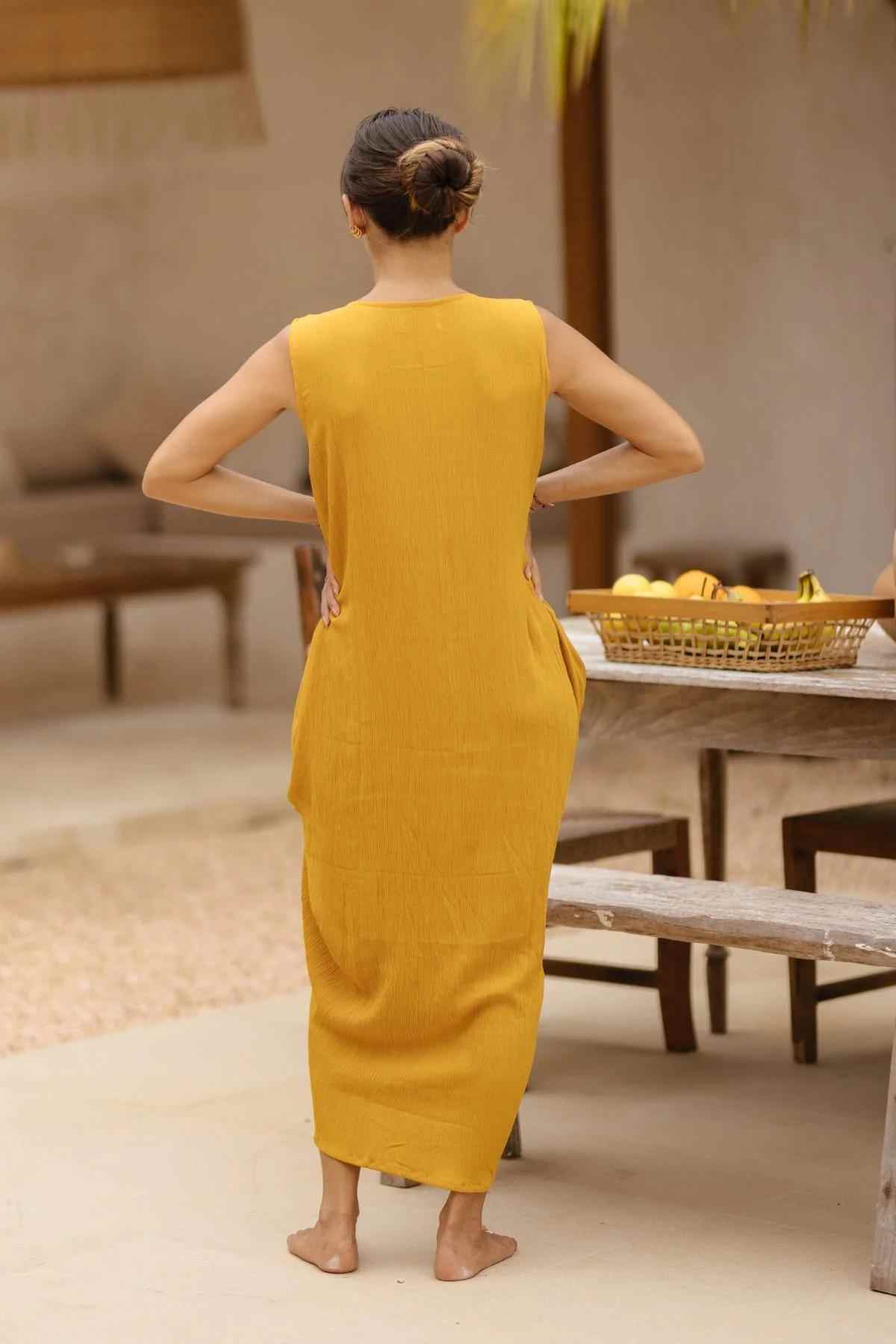 A person with dark hair in a bun stands barefoot with hands on hips, facing away from the camera. Clad in Myrah Penaloza's Virgo Luna Dress, featuring a sleeveless style and high neckline, they exude grace. Nearby is a wooden table holding a bowl of fruit. The setting appears to be an outdoor space with rustic decor.