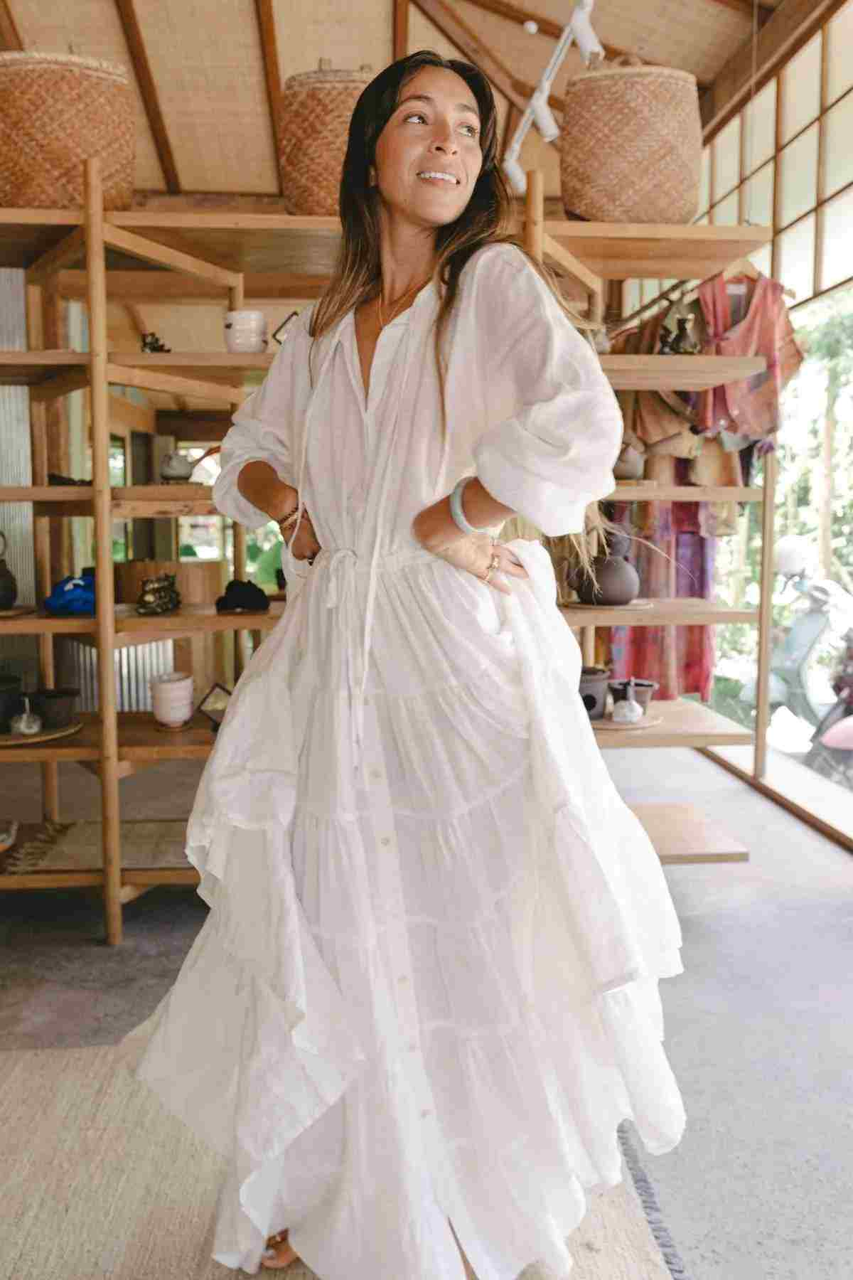 A woman wearing the La Mexicana Gown from Myrah Penaloza, featuring a flowing linen design with an adjustable waist, stands smiling in a bright room with her hands on her hips. Surrounded by wooden shelves and woven baskets, natural light pours in through large windows, highlighting lush plants outside. Her long hair cascades gracefully down her back.