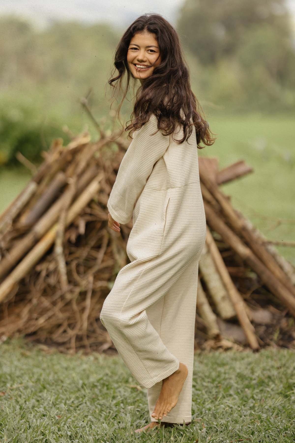 A woman with long, dark hair is smiling and looking back over her shoulder while standing barefoot on grass, wearing a light-colored Camper Playsuit by Myrah Penaloza. In the background, a pile of logs and branches hints at adventurous weekend getaways.